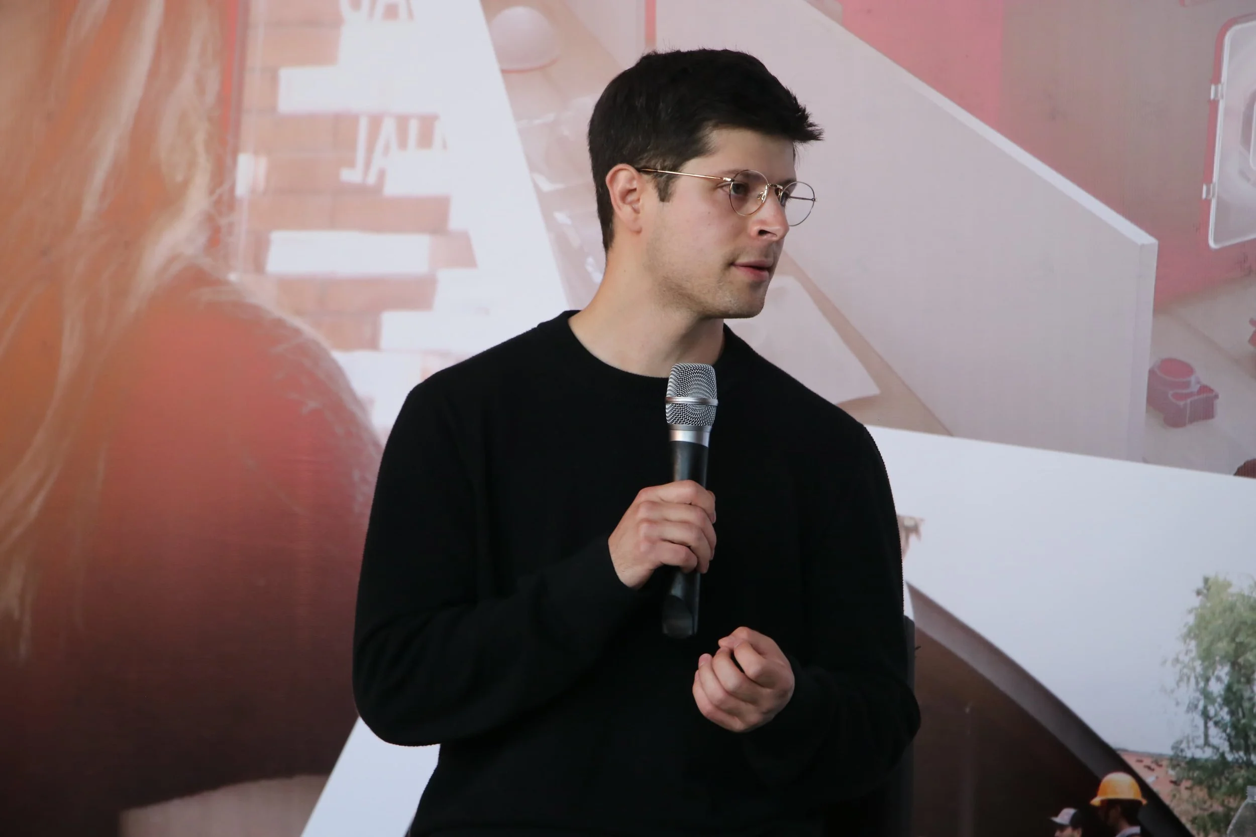 Hombre joven con gafas y sudadera negra sosteniendo un micrófono en un entorno interior, con fondo de decoraciones y objetos en tonos pastel.