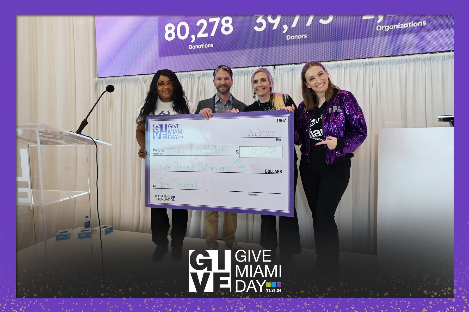 Group of four people holding a large ceremonial check at a charity event, with a purple and white backdrop displaying donation numbers, celebrating Give Miami Day. The check is made out for one thousand dollars to Fina High 4 Haiti, signed and dated