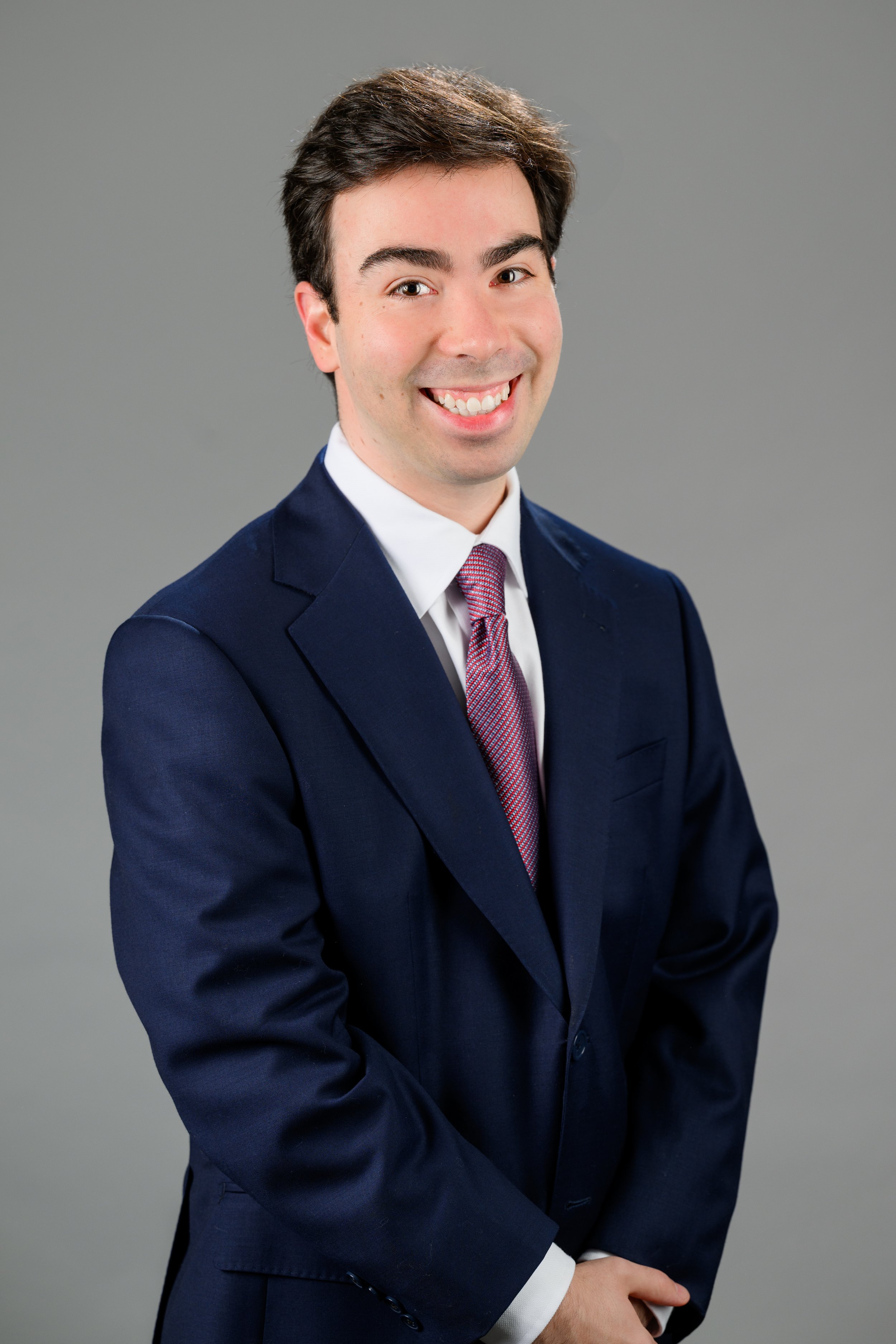 A young man in a dark blue suit, white shirt, and pink tie smiling and posing with hands clasped in front against a gray background.