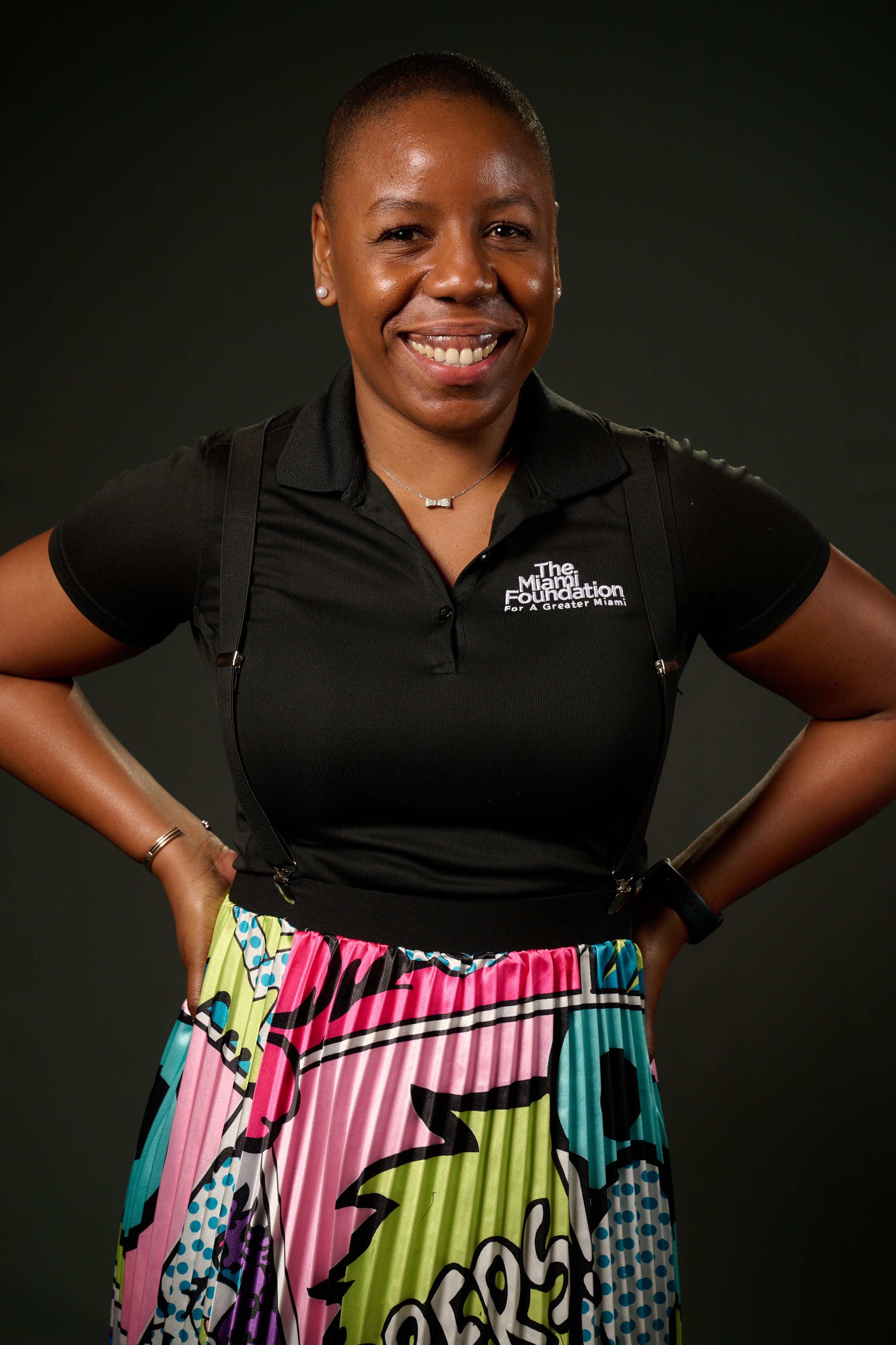 A smiling woman with short hair, wearing a black polo shirt with the logo 'The Miami Foundation' and a colorful, comic book style skirt. She has her hands on her hips and stands against a dark background.
