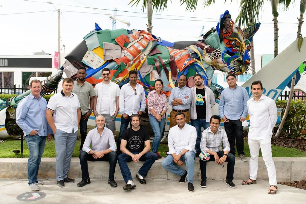 Group of people posing in front of a large, colorful, abstract sculpture in an outdoor setting with palm trees.