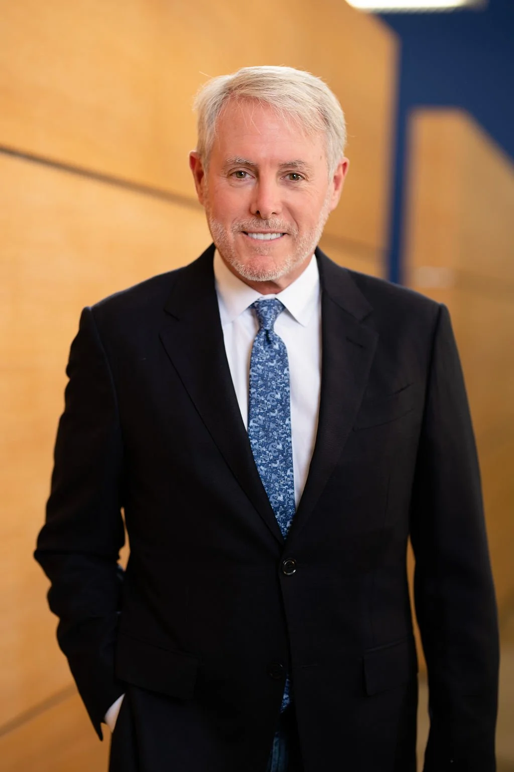 Man in a suit with a blue patterned tie standing indoors, smiling.