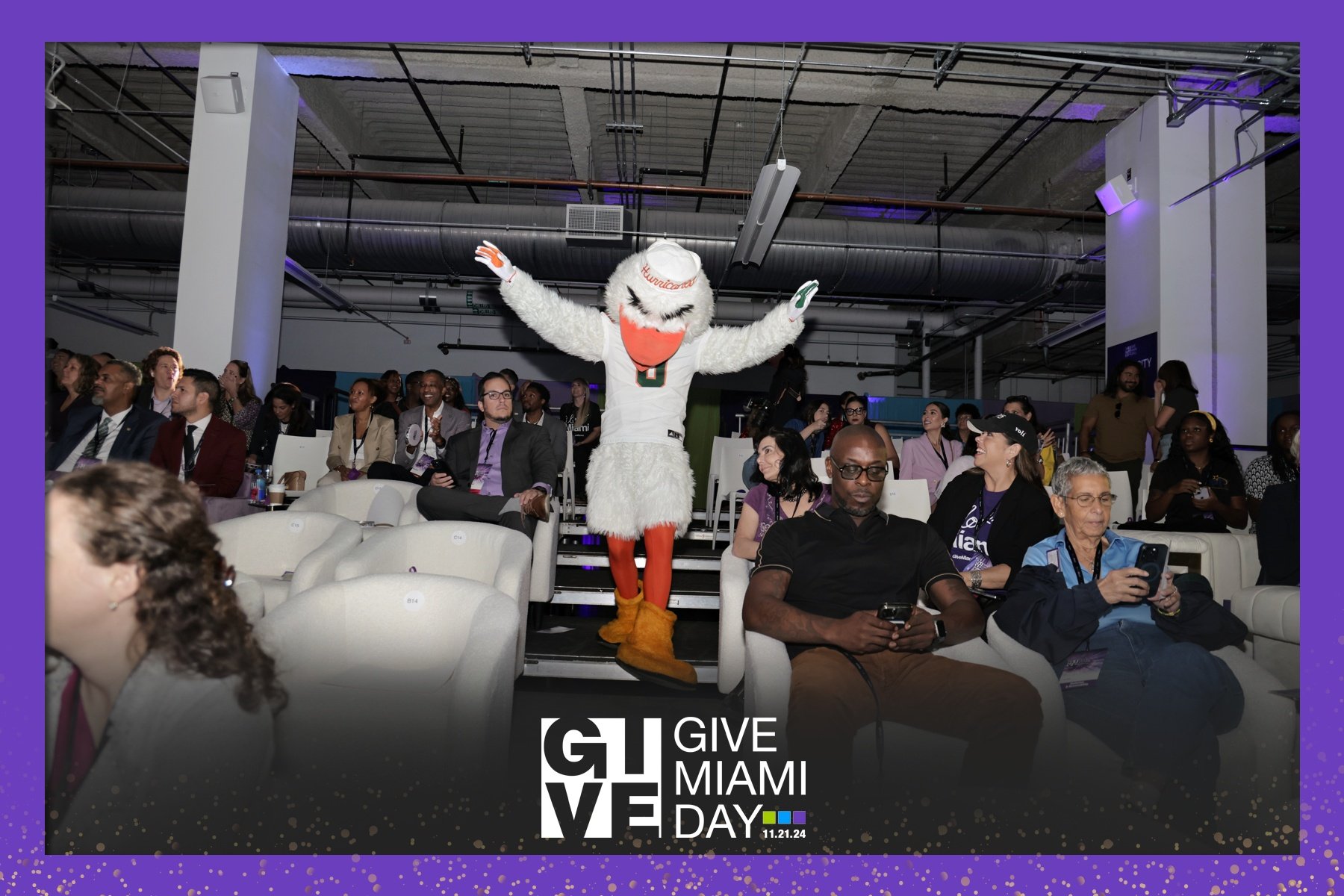 An indoor conference with a diverse group of attendees seated, some using phones, and a person in an all-white bird mascot costume with outstretched arms walking down the aisle. The event is called 'Give Miami Day' on November 21, 2024, with branding