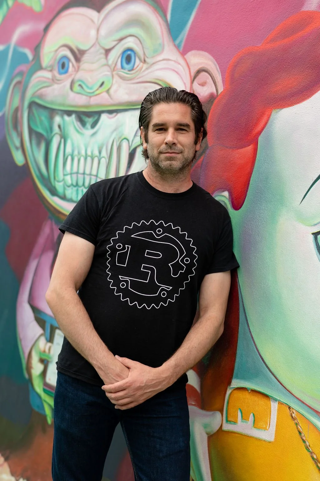 Man in a black T-shirt with a Rust logo standing in front of a colorful mural featuring large cartoonish faces.
