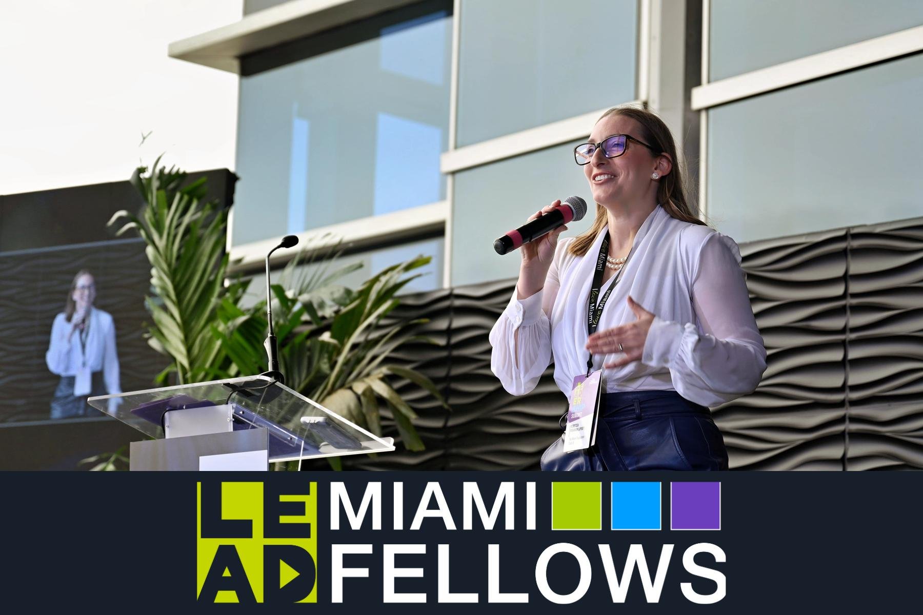 A woman wearing glasses, a white blouse, and a pink necklace speaks into a microphone at an outdoor event, with a backdrop displaying the Miami Ad Fellows logo.