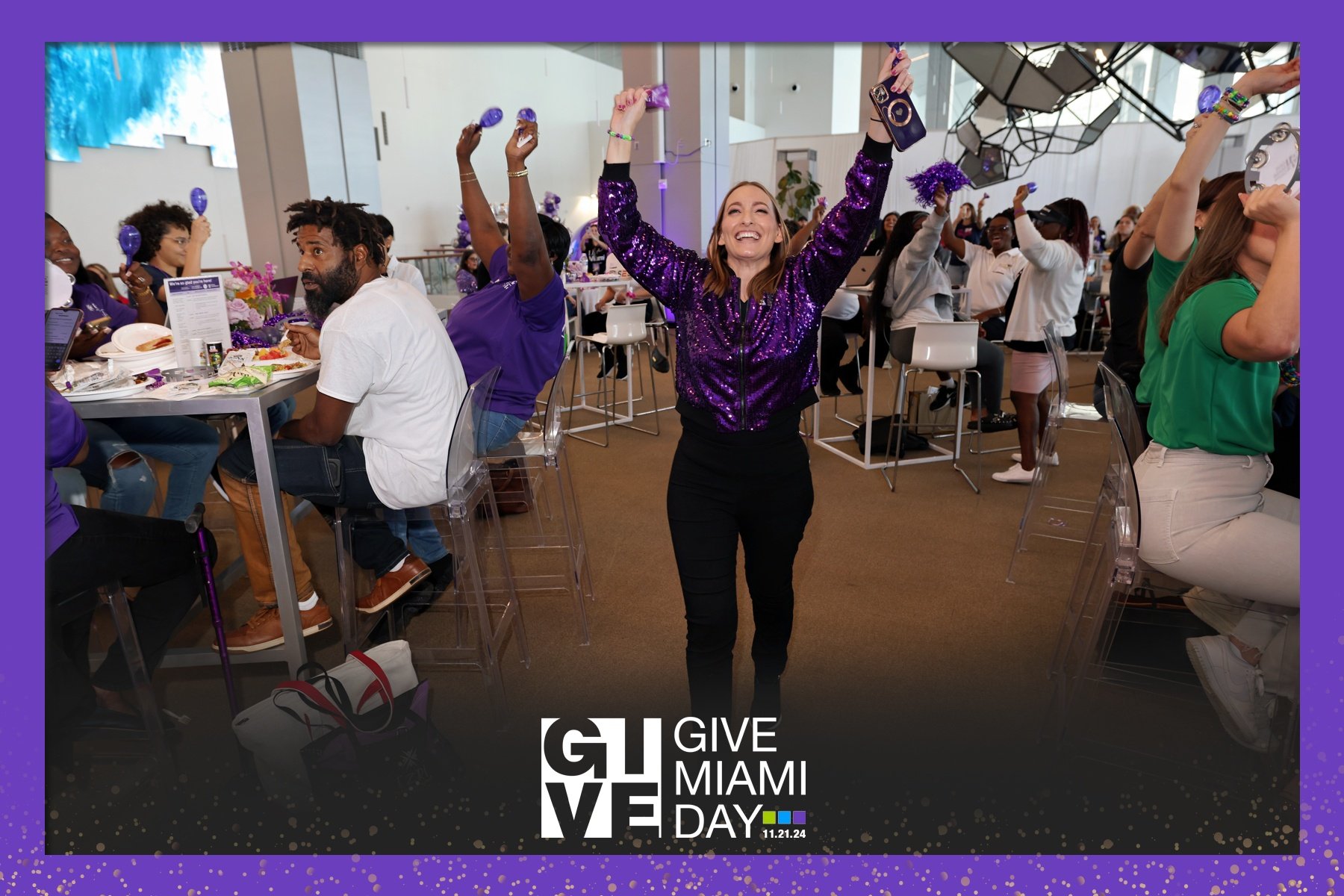 A woman in a purple sequined jacket smiling and raising her arms in celebration at Give Miami Day event, surrounded by people at tables with purple decorations and snacks.