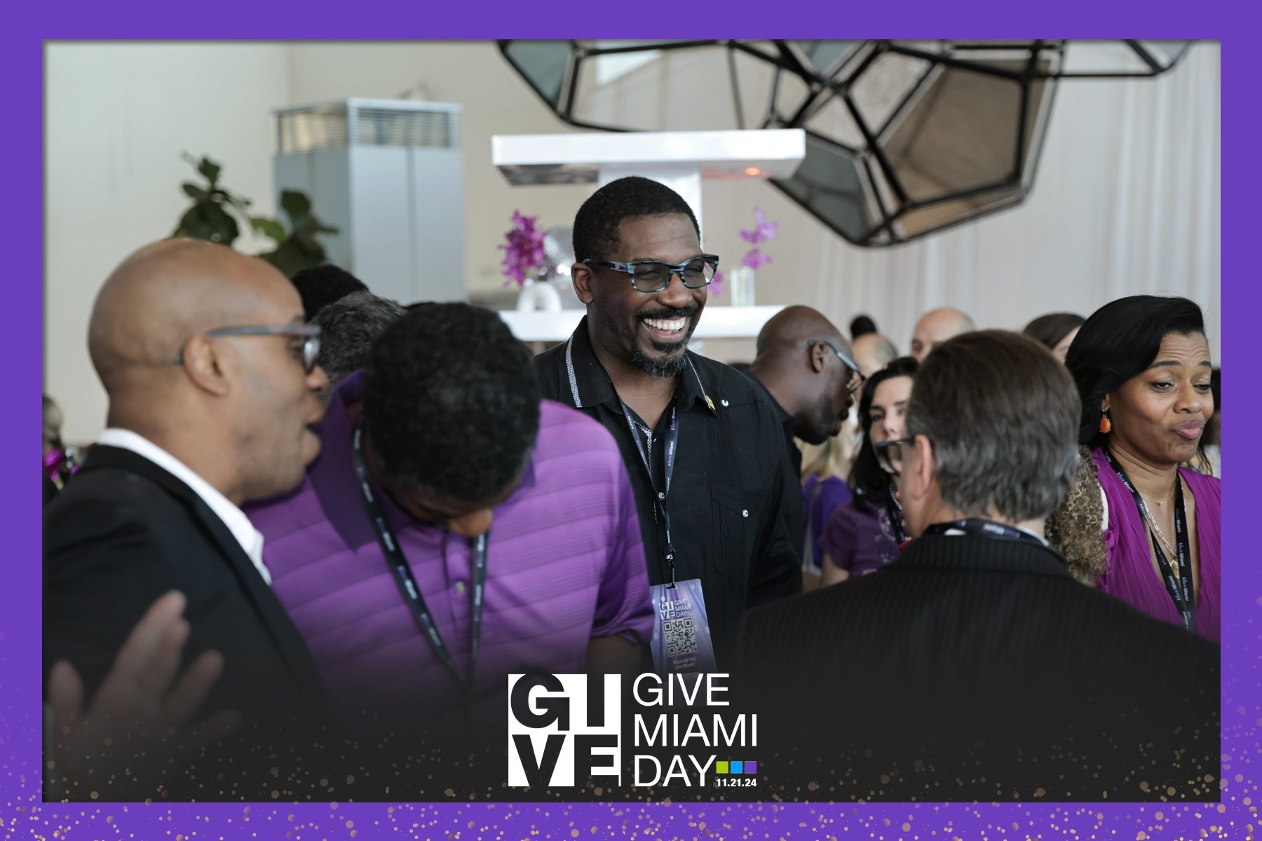 Group of people at Give Miami Day event, smiling and conversing, with a man in glasses in the center. There is a purple border with gold dots and the event logo at the bottom.