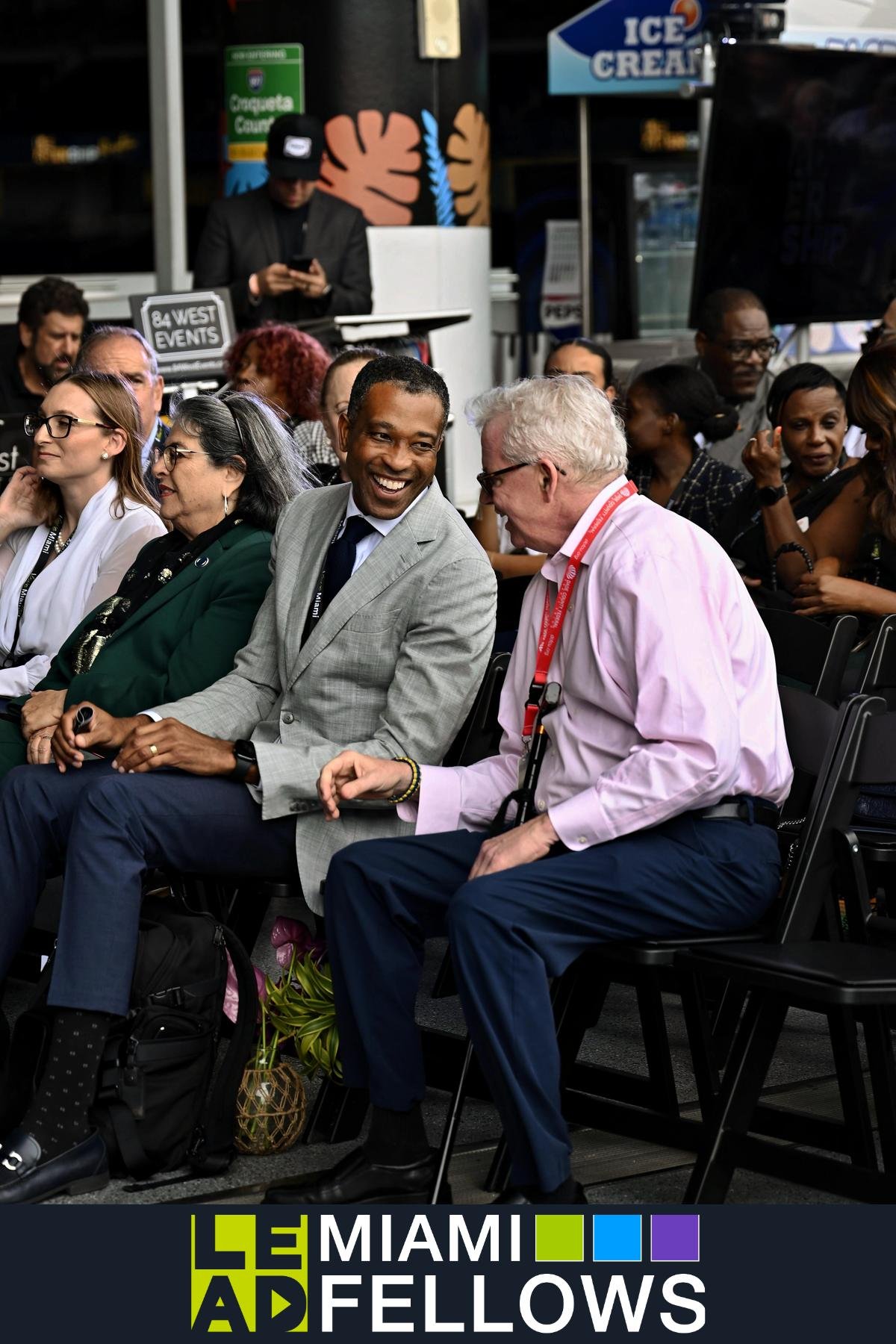 Audience at Miami Delays event, two men in the foreground are talking and smiling, surrounded by diverse attendees seated in rows.