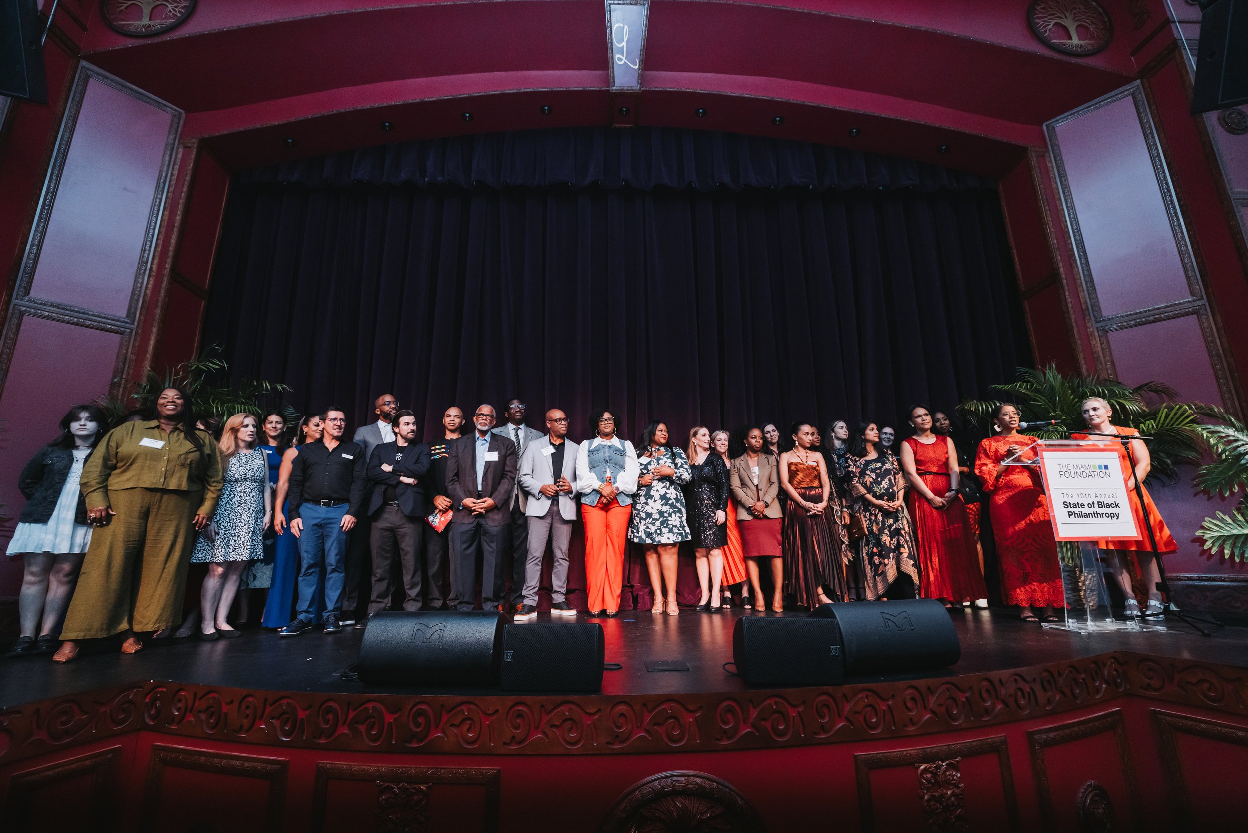 Group of diverse people standing on stage at the State of Black Philanthropy event, with a speaker at a podium labeled 'The Miami Foundation' and 'The 10th Annual State of Black Philanthropy' in an ornate theater with red walls and black curtains.
