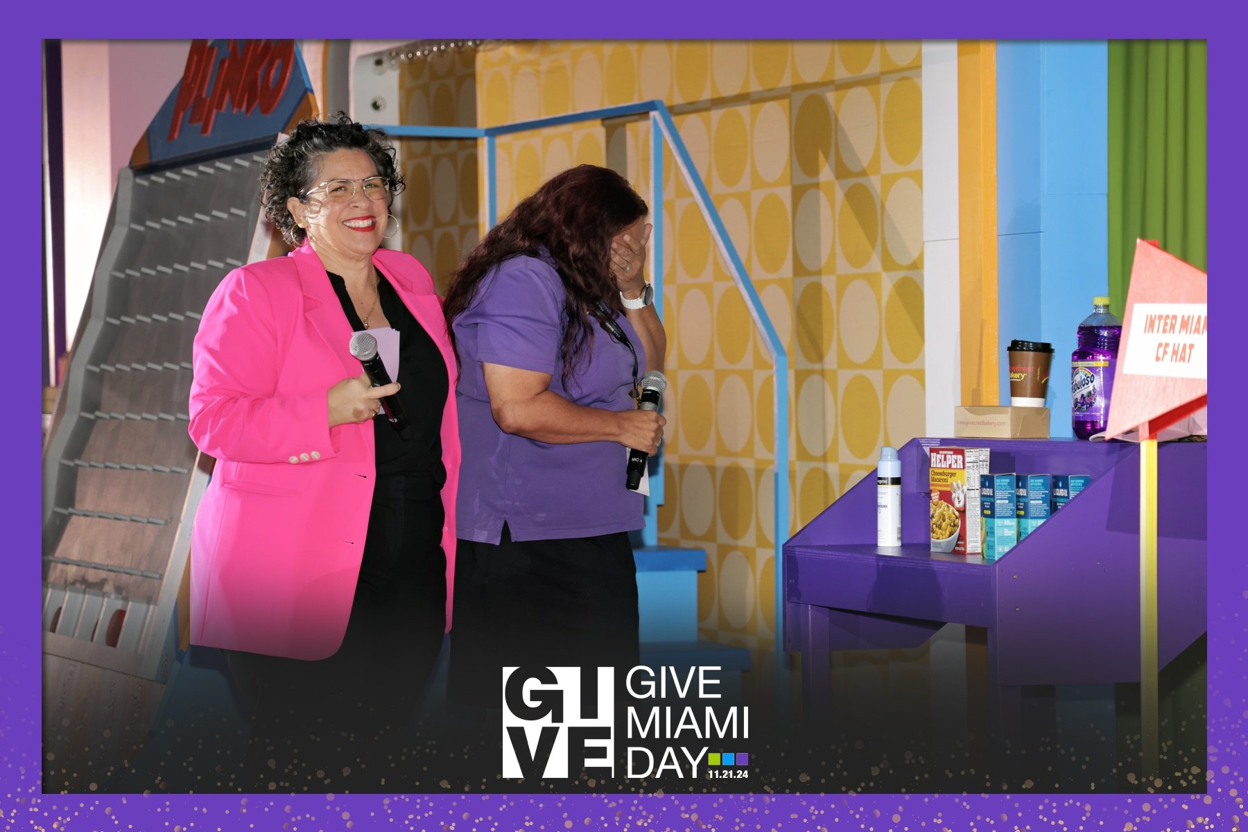 Two women with microphones standing near a snack stand, one smiling and the other holding her head in distress, during Give Miami Day event.