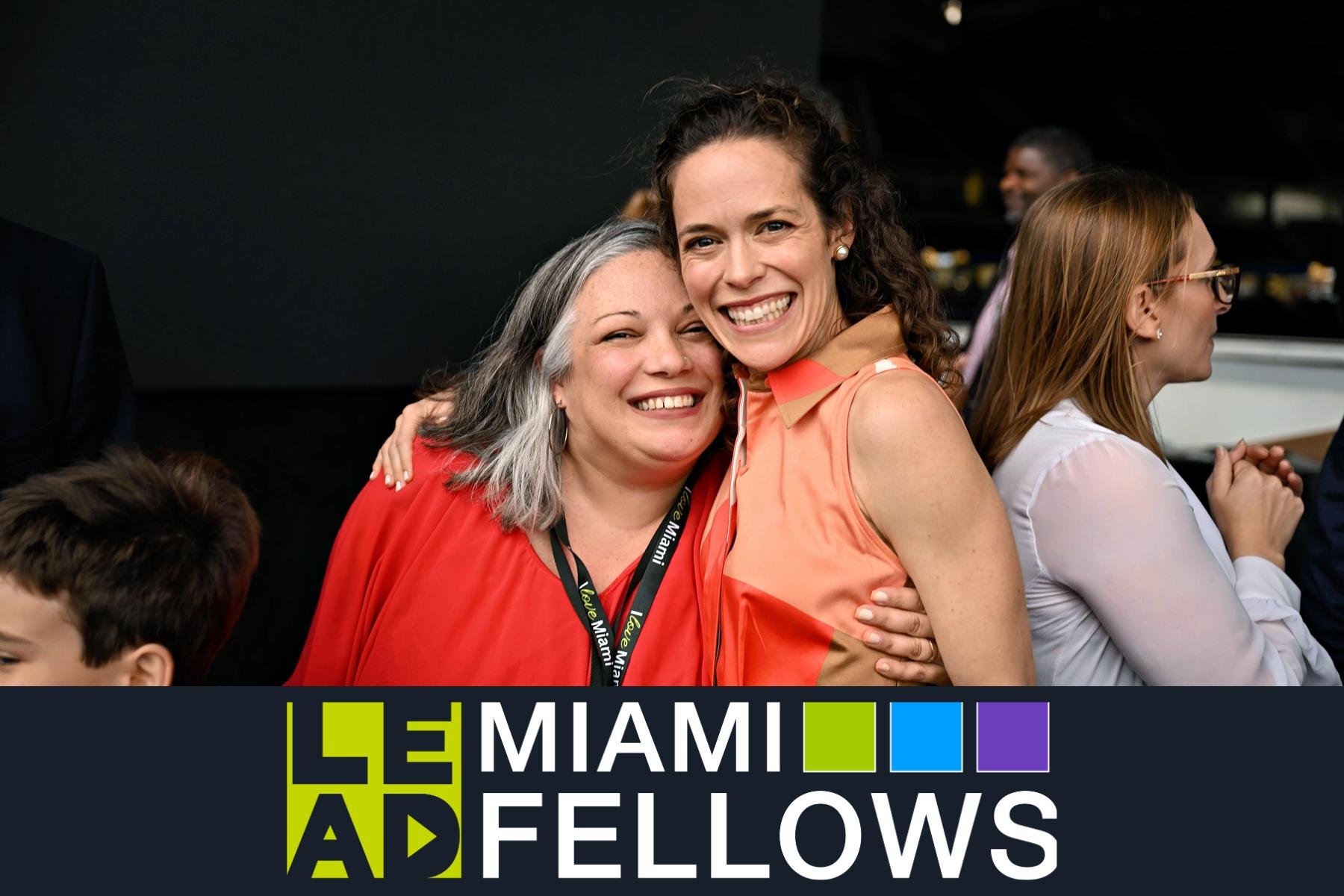 Two women hugging and smiling, at an event with a Miami Miami Fellows logo at the bottom.