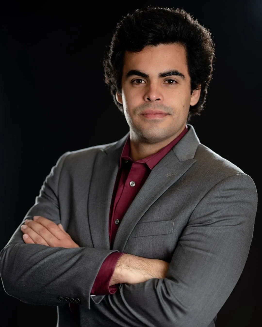 Man in a grey suit with burgundy shirt, arms crossed, against a dark background.