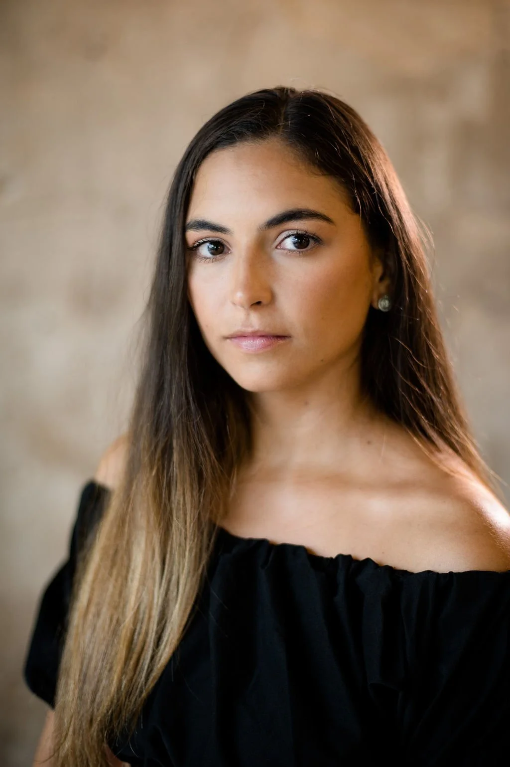 A woman with long hair wearing a black off-the-shoulder top, photographed against a neutral background.