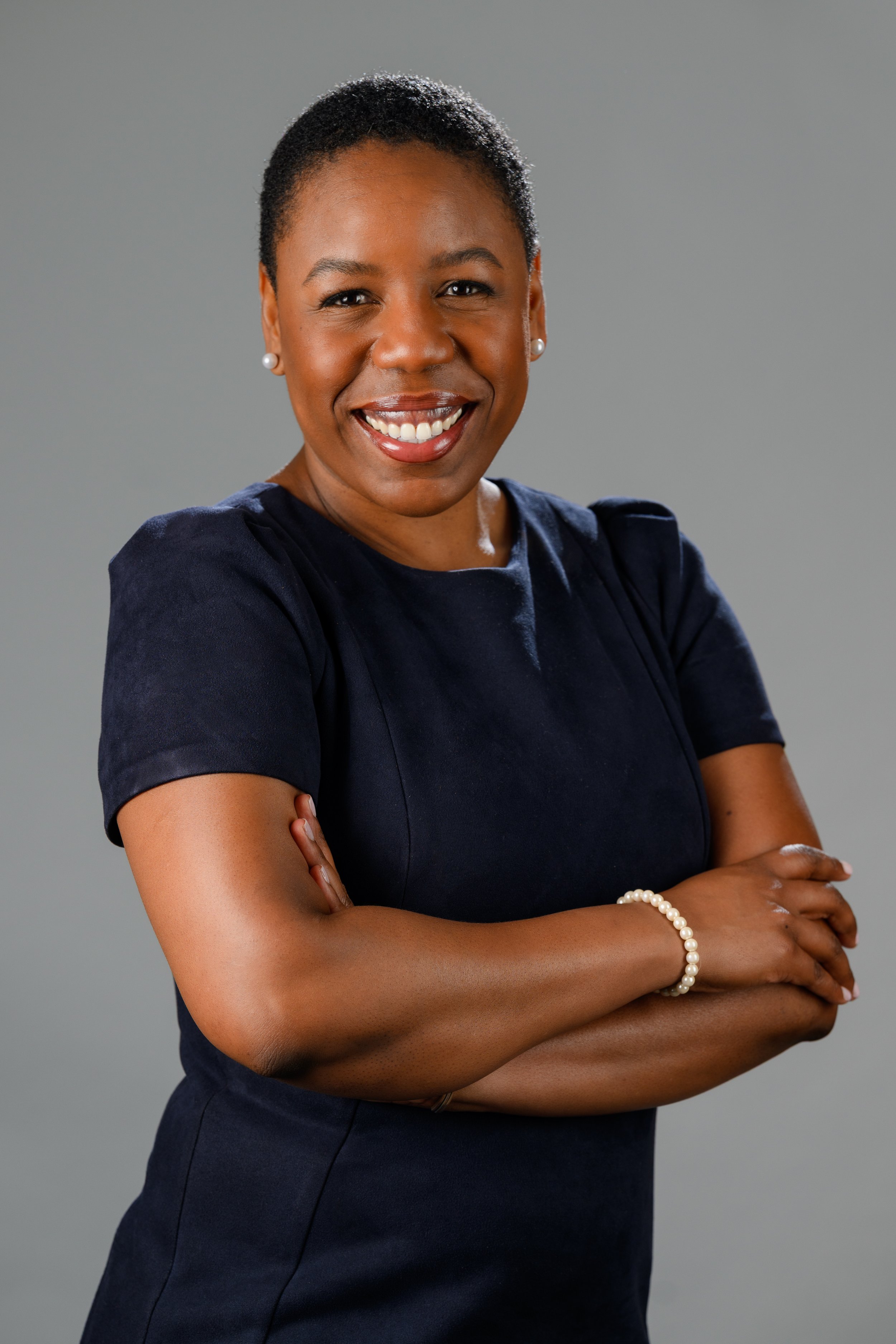 A smiling African American woman with short hair, crossed arms, wearing a navy blue dress, pearl earrings, and a pearl bracelet against a gray background.