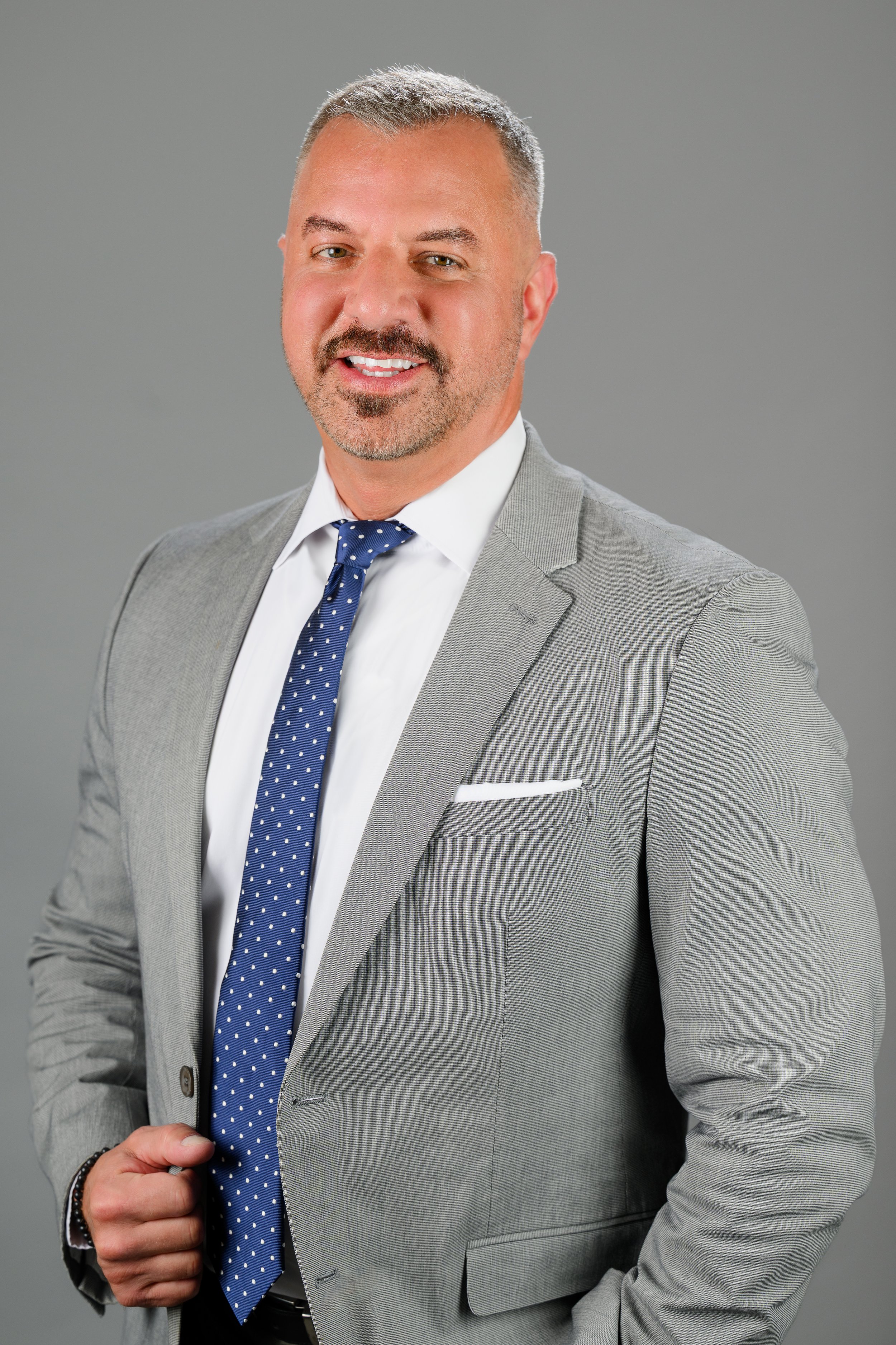 A man wearing a gray suit, white shirt, and blue polka dot tie, smiling, against a gray background.