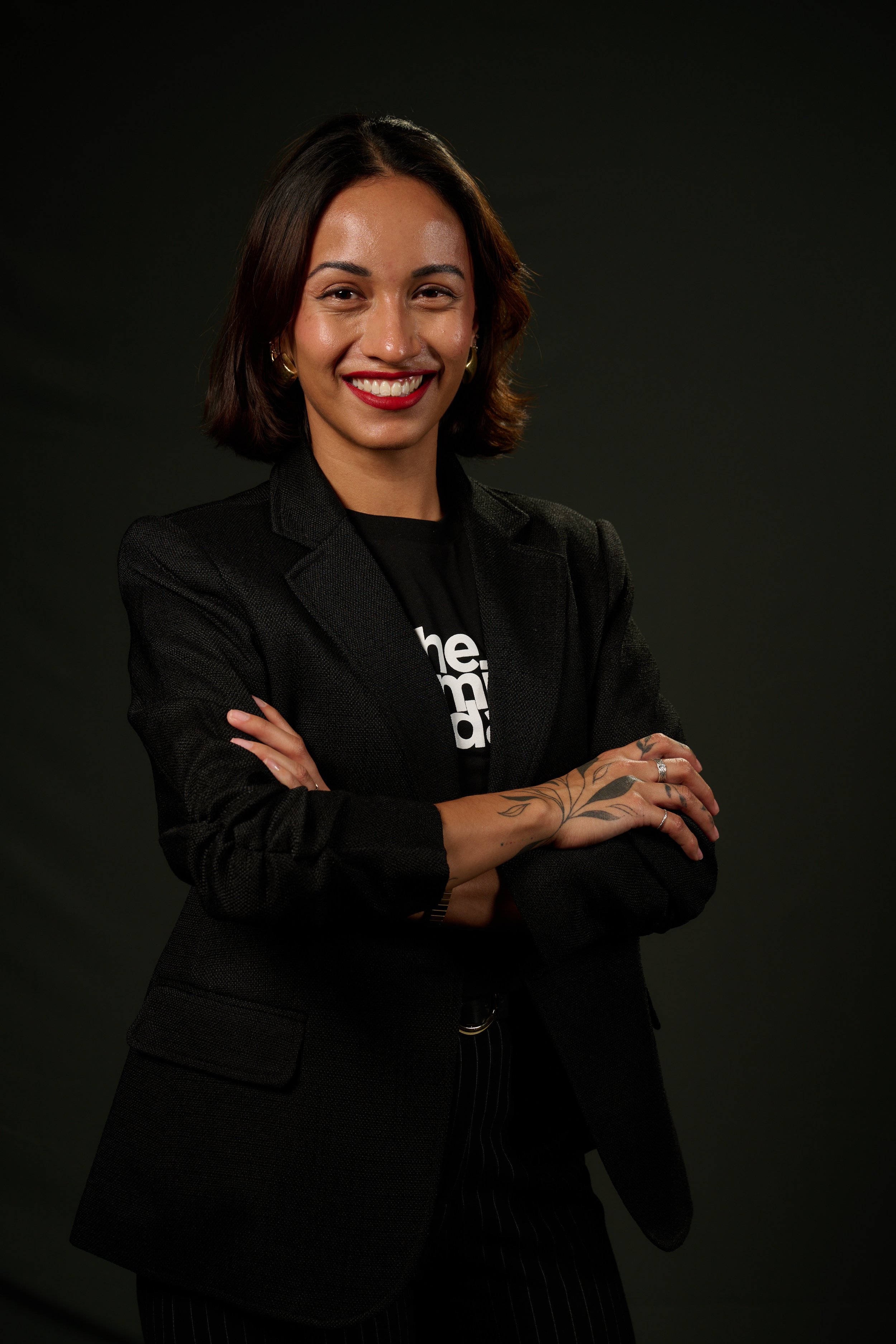 A woman with shoulder-length dark hair, wearing a black blazer and a black t-shirt, smiling with her arms crossed, against a dark background.
