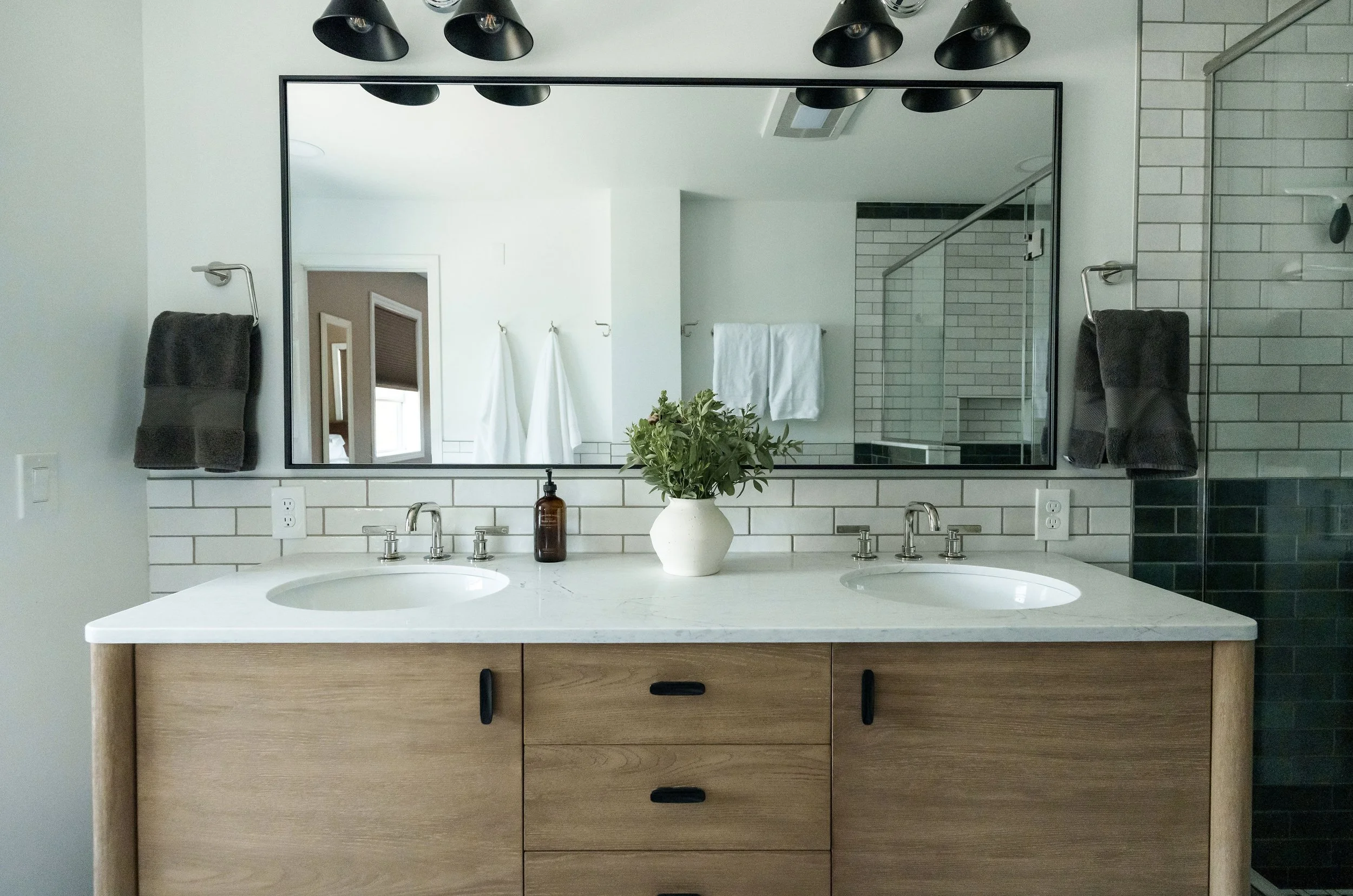 Bathroom vanity with two sinks, a large mirror, dark towels on wall hooks, a potted plant, and a shower area with white subway tiles and a glass door.