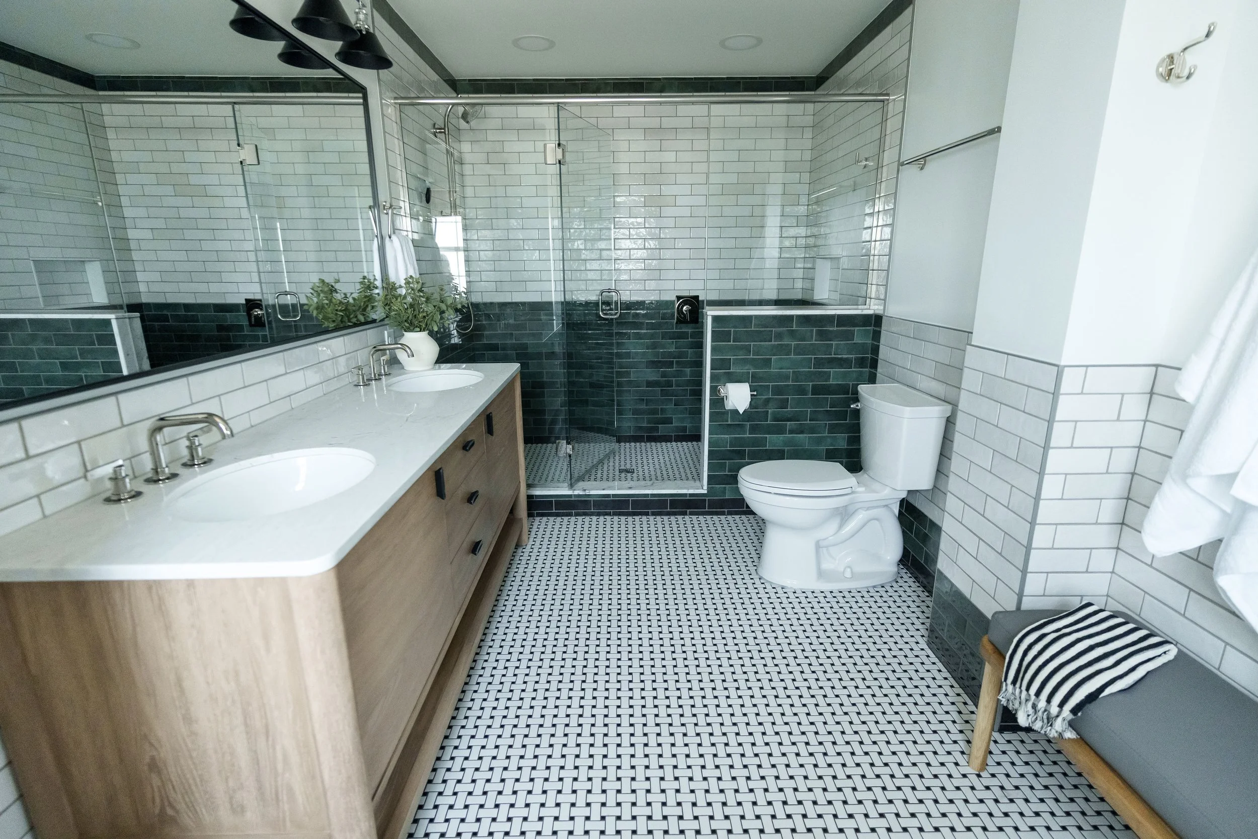 Modern bathroom with a double vanity with a marble countertop, a large mirror, a glass-enclosed shower, a toilet, a wooden bench with a striped towel, and tiled floors and walls in shades of white, gray, and dark green.