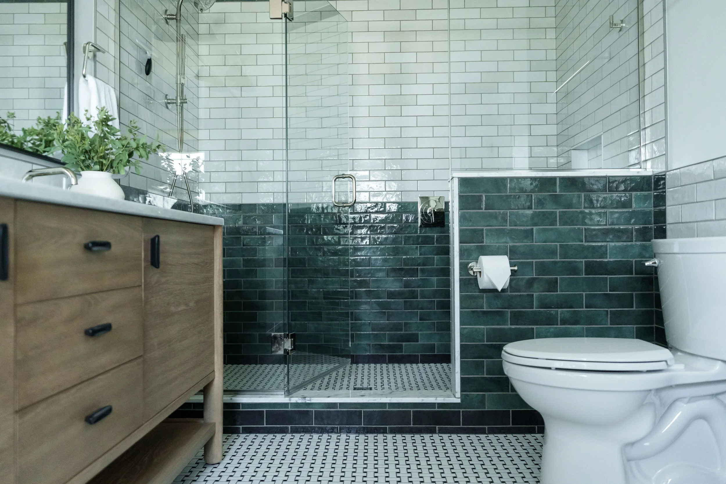 Modern bathroom with glass shower enclosure, white and dark green subway tiles, toilet, wooden vanity, and black and white patterned floor.