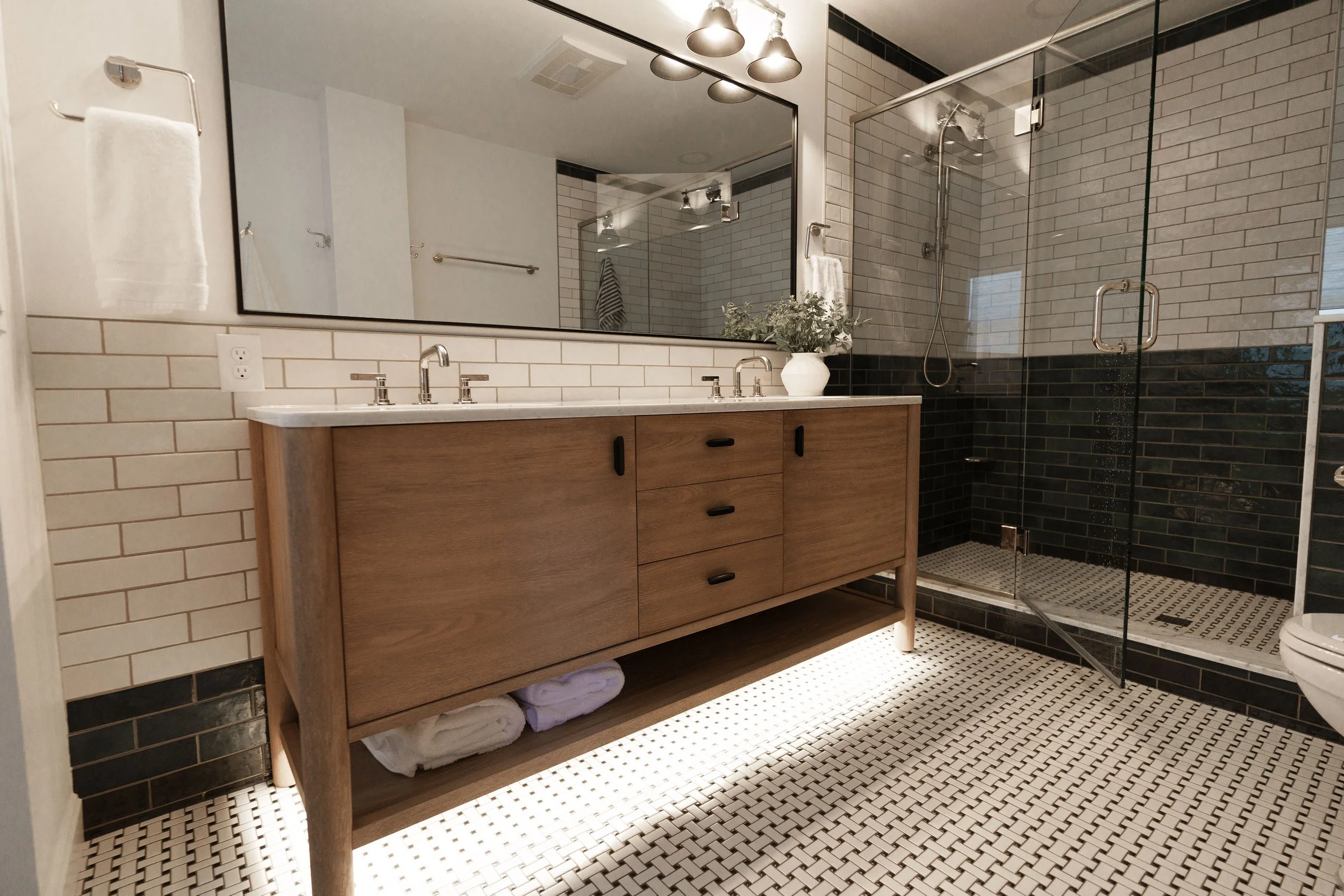 Modern bathroom with wooden vanity, large mirror, glass shower enclosure, black and white tile floor.