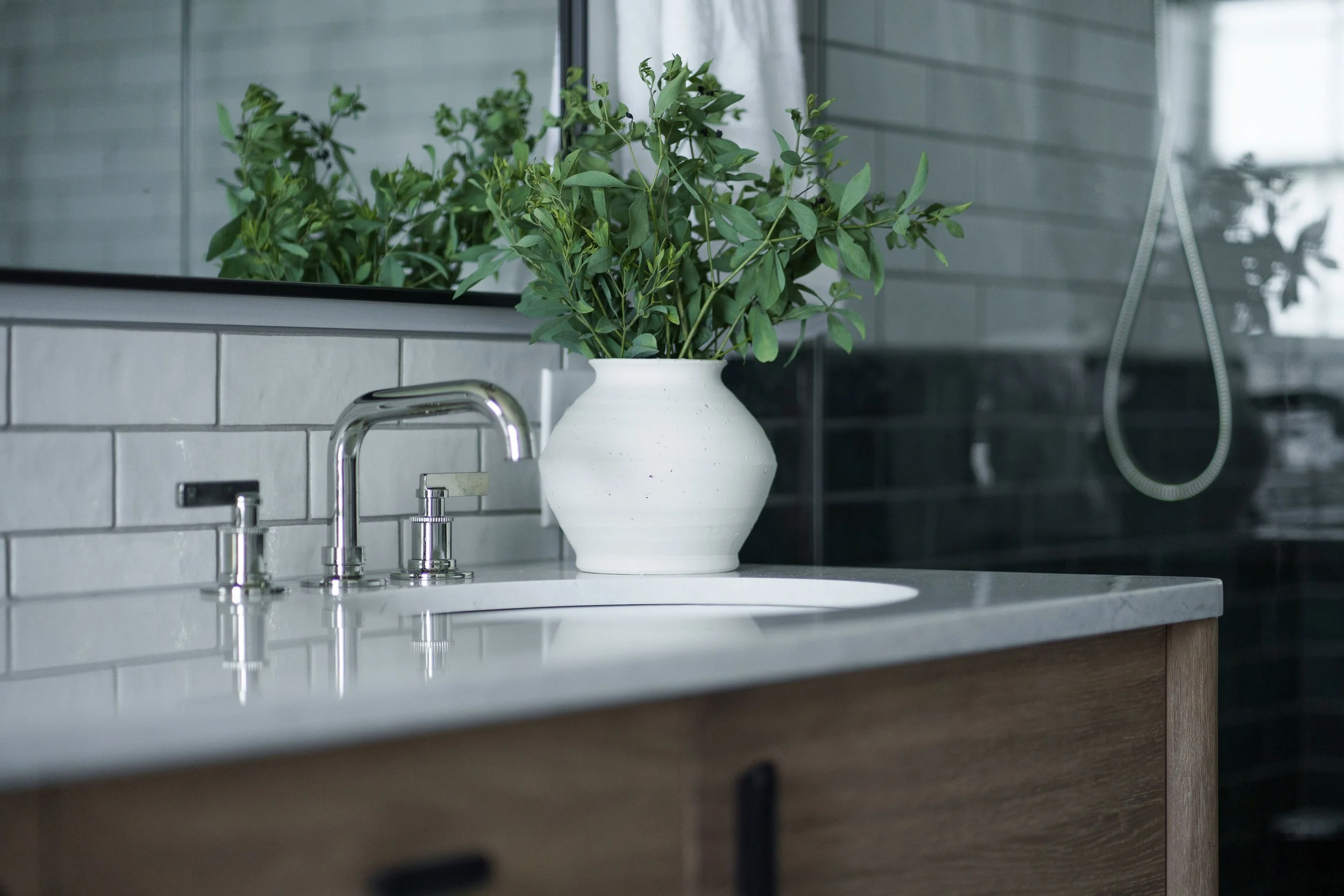 White bathroom sink with a faucet and a large green plant in a white vase on the countertop, with a mirror and tiled wall behind.