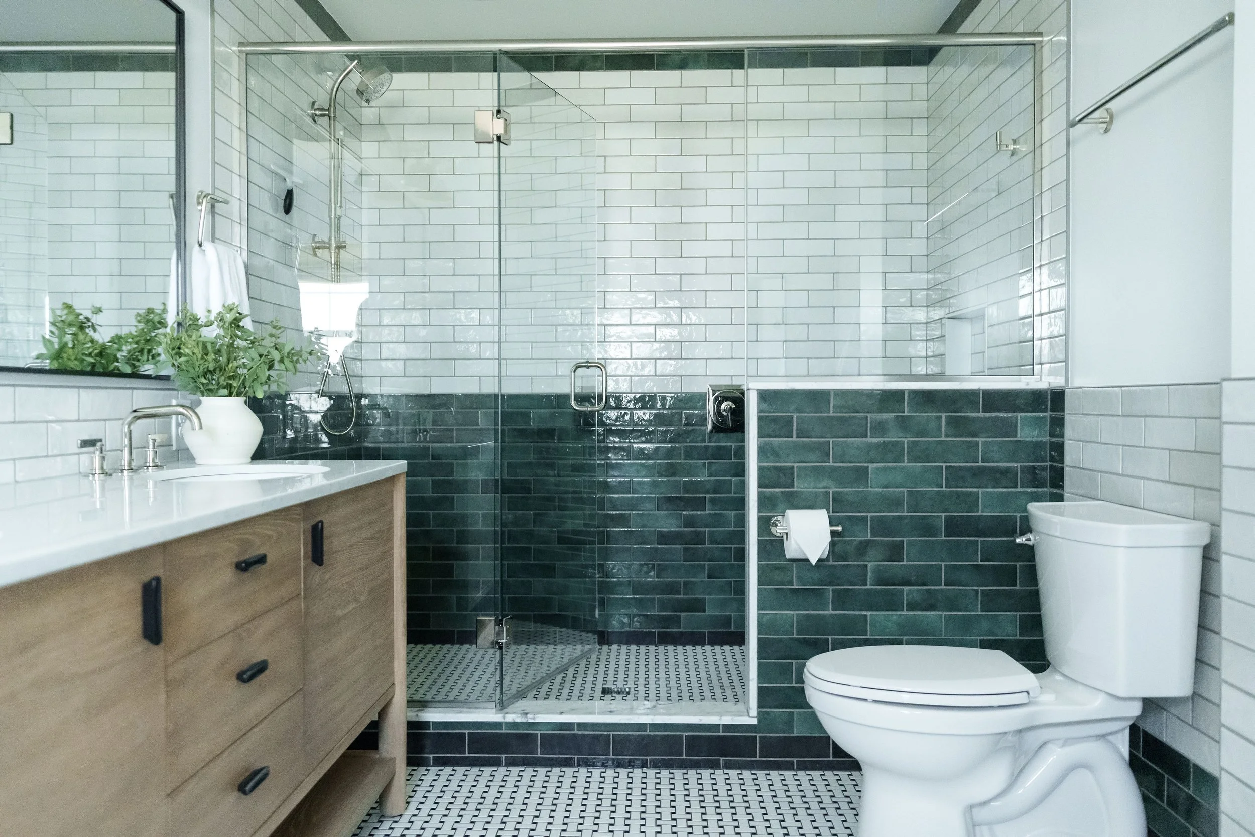 Modern bathroom with glass-enclosed shower, white and dark green subway tiles, wooden vanity with a white countertop, potted plant, and a white toilet.