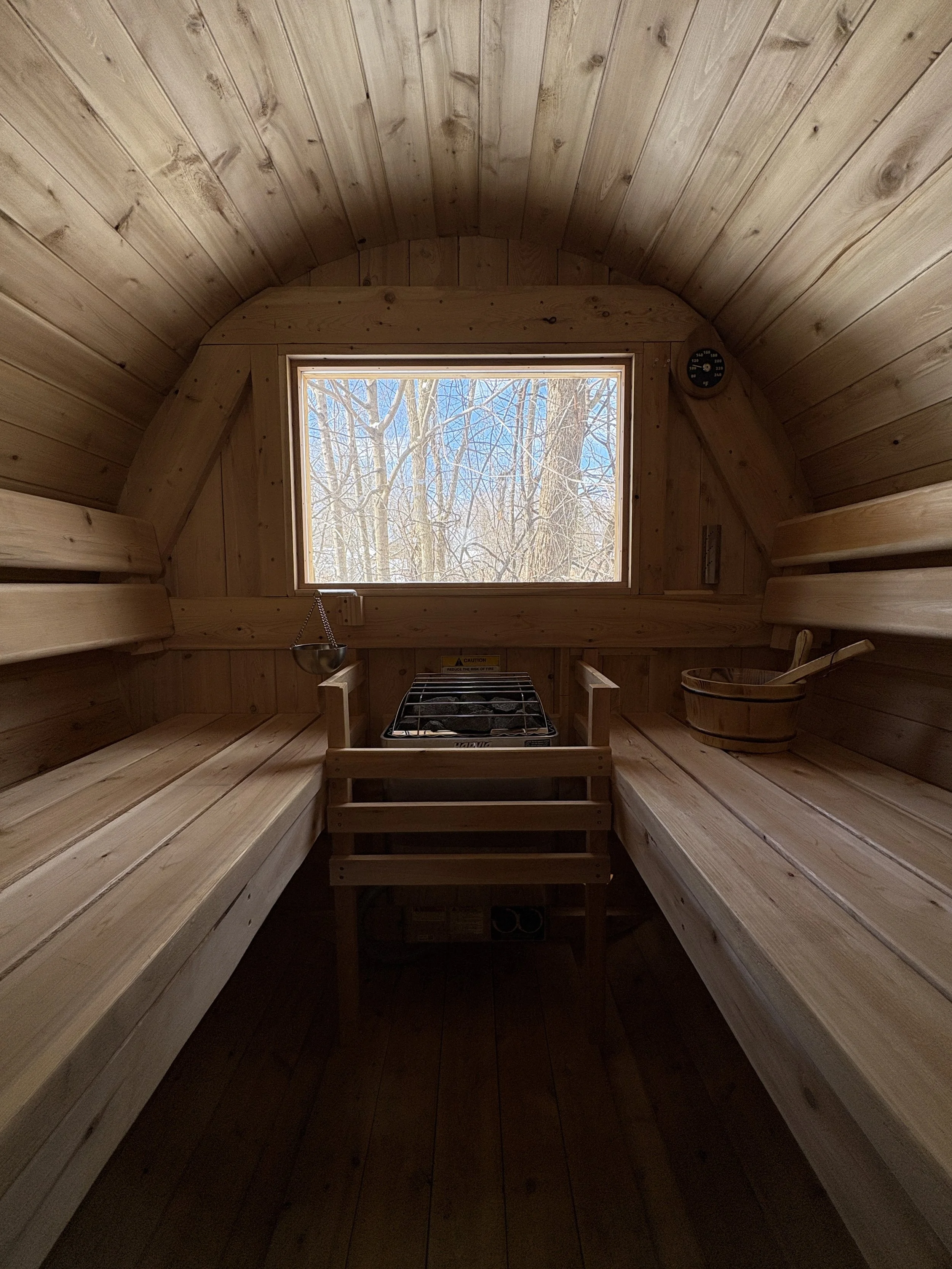 Interior of a small wooden sauna with benches on both sides, a window showing trees outside, a small stove in the center, a wooden bucket, and a ladle on the right bench.