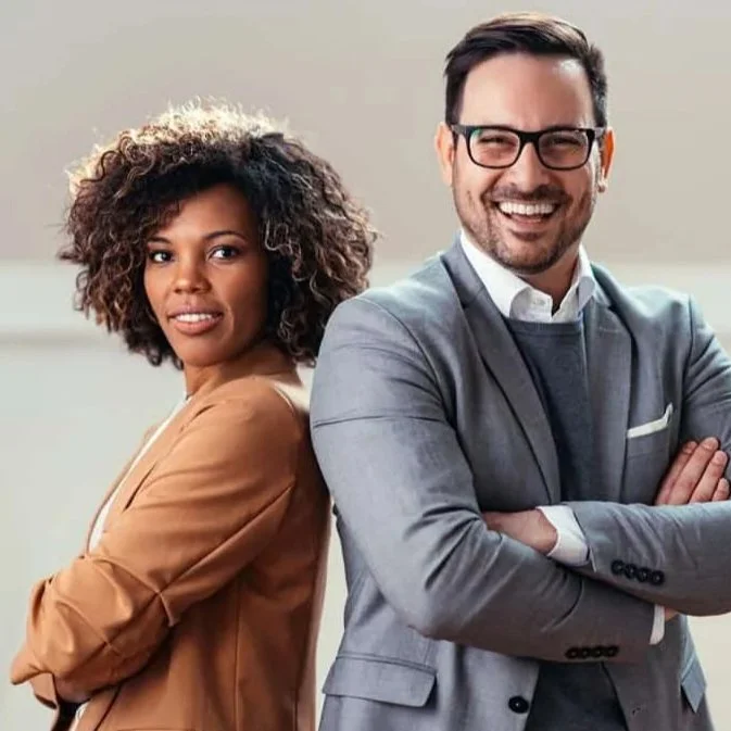 A man and a woman stand back-to-back in a professional setting, smiling confidently. The woman has curly hair and is wearing a tan blazer, while the man has short dark hair, glasses, and is dressed in a gray suit with a white shirt.
