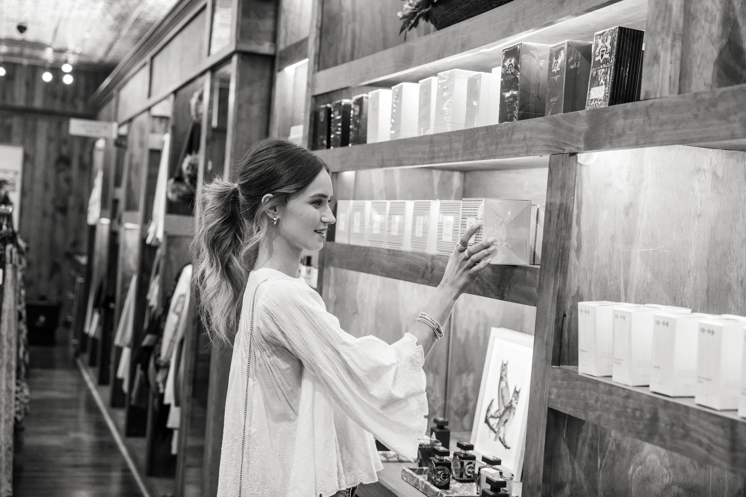A woman browsing books and items on a wooden shelf in a store.
