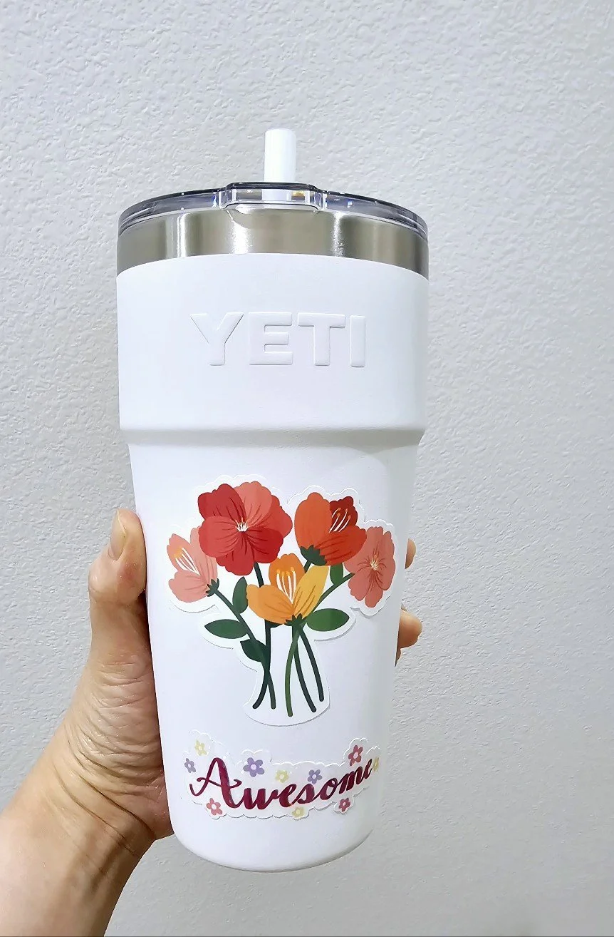 A white YETI tumbler with a floral design and the word 'Awesome' written on it, held in front of a plain wall.