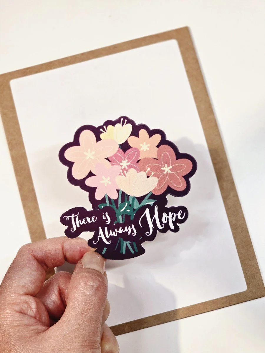 A hand holding a sticker of pink and cream flowers with the text "There is Always Hope" on it.