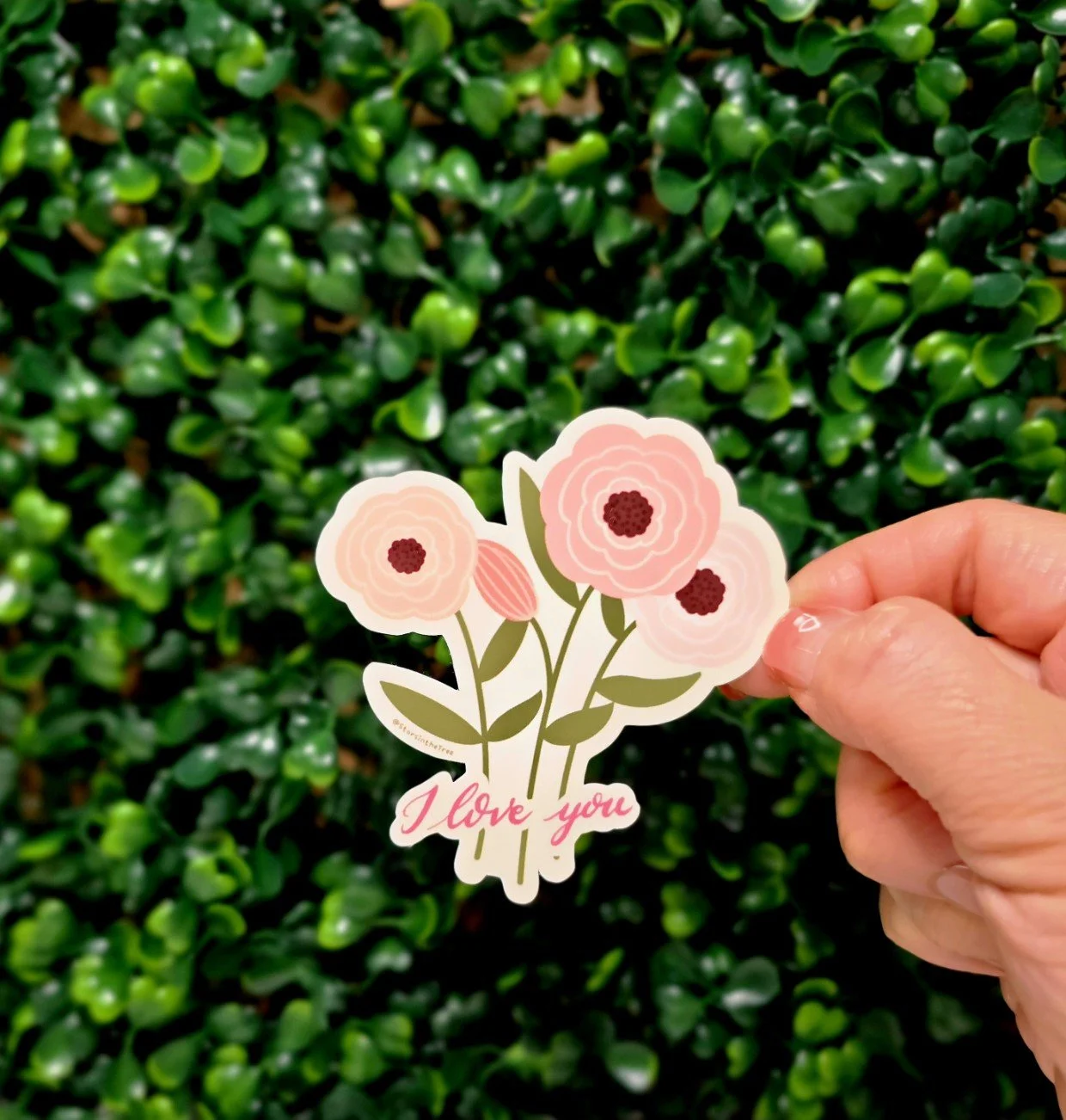 A hand holding a sticker with pink flowers and the words 'I love you' in front of a green leafy background.