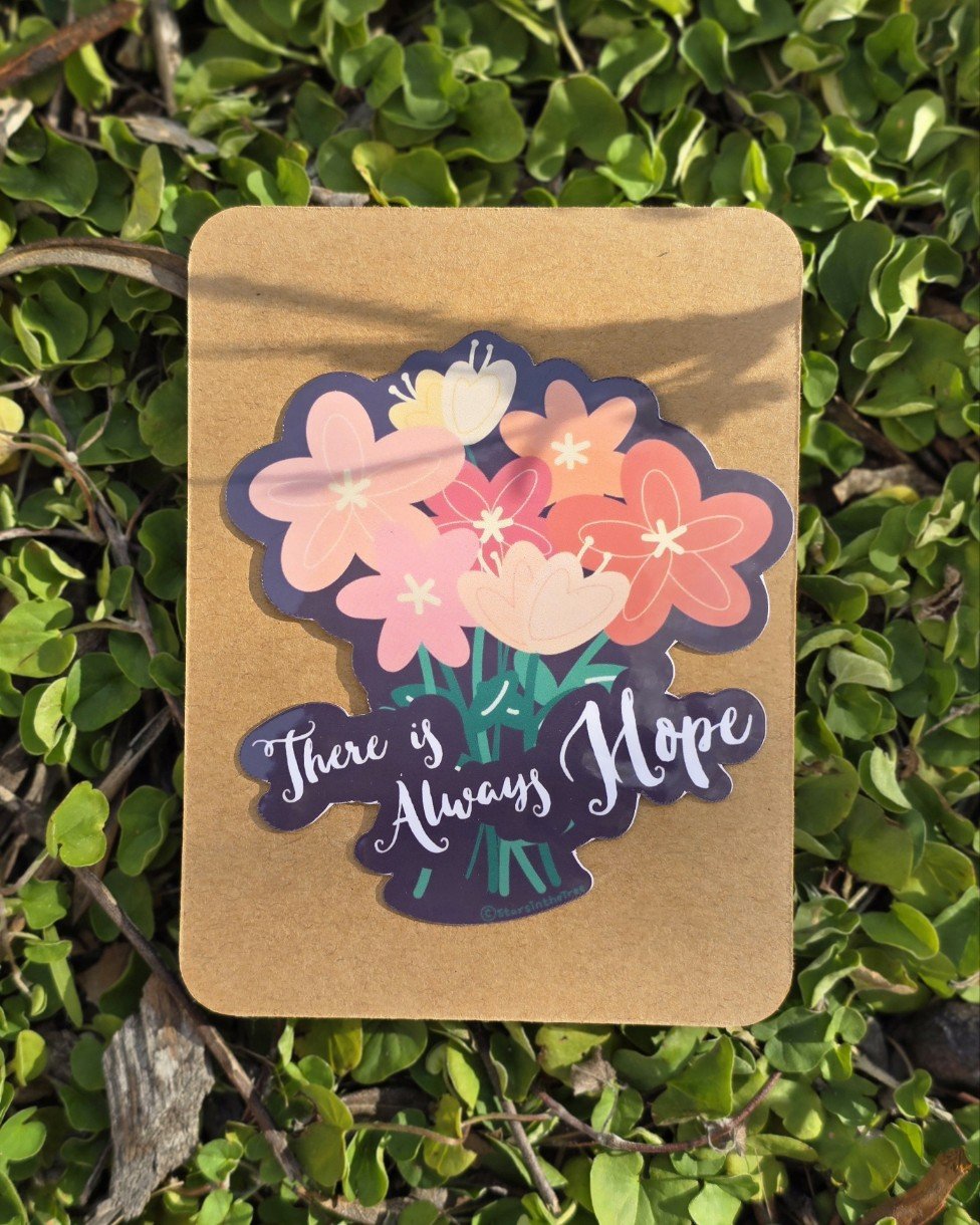Colorful sticker of a bouquet of flowers with pink, peach, and yellow blooms, and the phrase 'There is Always Hope' written across it, placed on a brown paper background among green leaves.