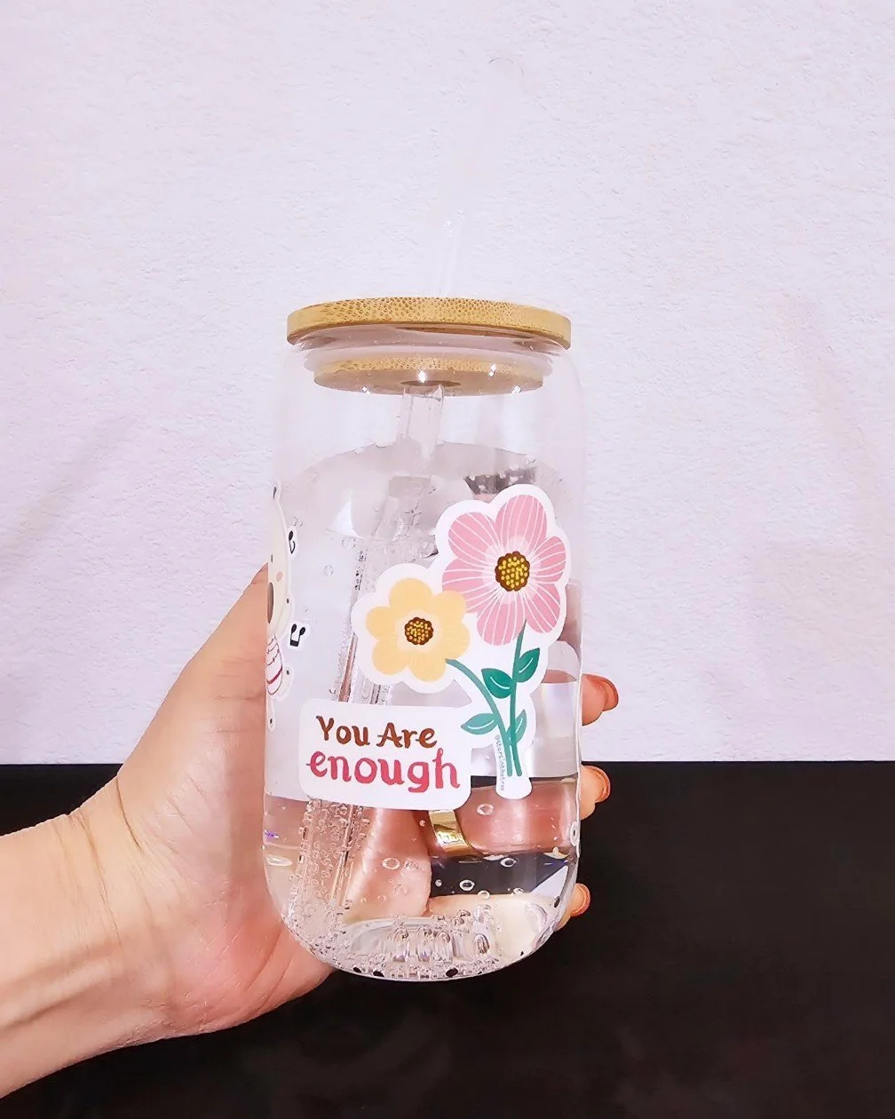 A hand holding a glass jar with a gold-colored lid, decorated with stickers of pink and yellow flowers and the phrase "You Are enough". The jar contains clear liquid and ice cubes, with a metal straw inside.
