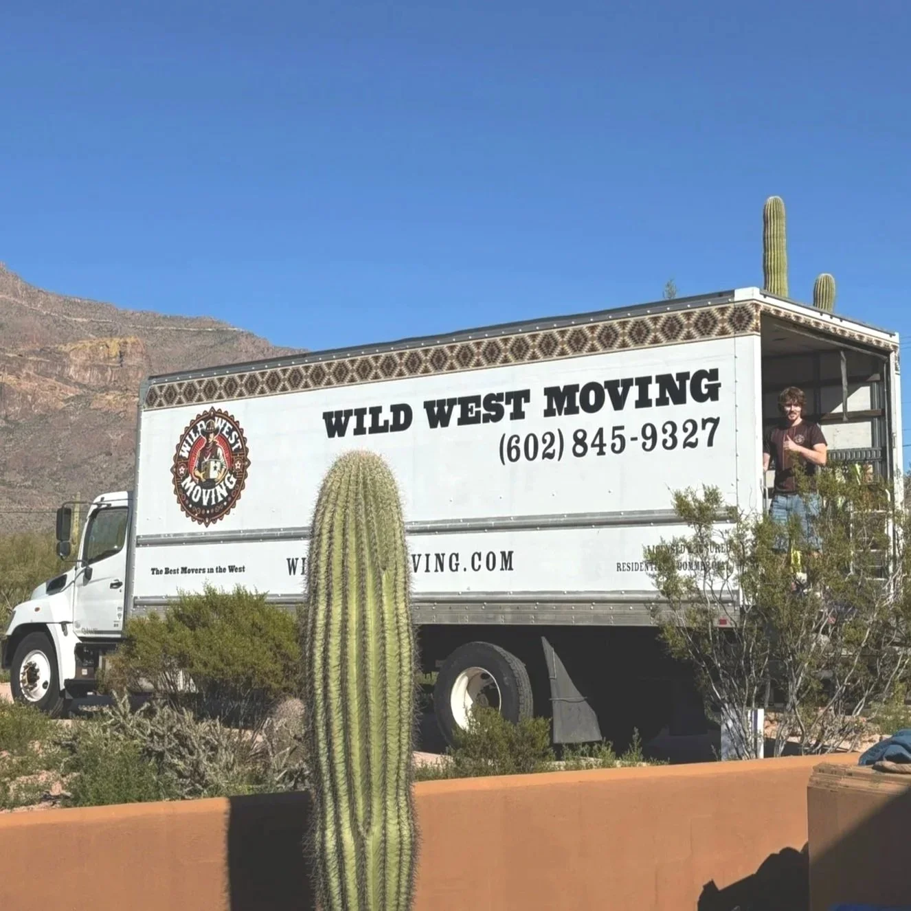 A moving truck with the company name Wild West Moving, phone number, and logo, parked outdoors with desert plants and cacti in front of a mountainous landscape under a blue sky.