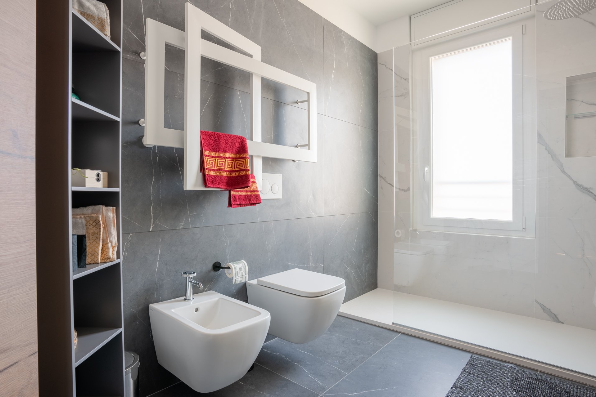 Bagno moderno con pareti e pavimenti in pietra grigia, finestra grande, doccia con parete di vetro, lavabo, WC e bidet bianchi, mensola con asciugamani, specchio sopra il lavabo.