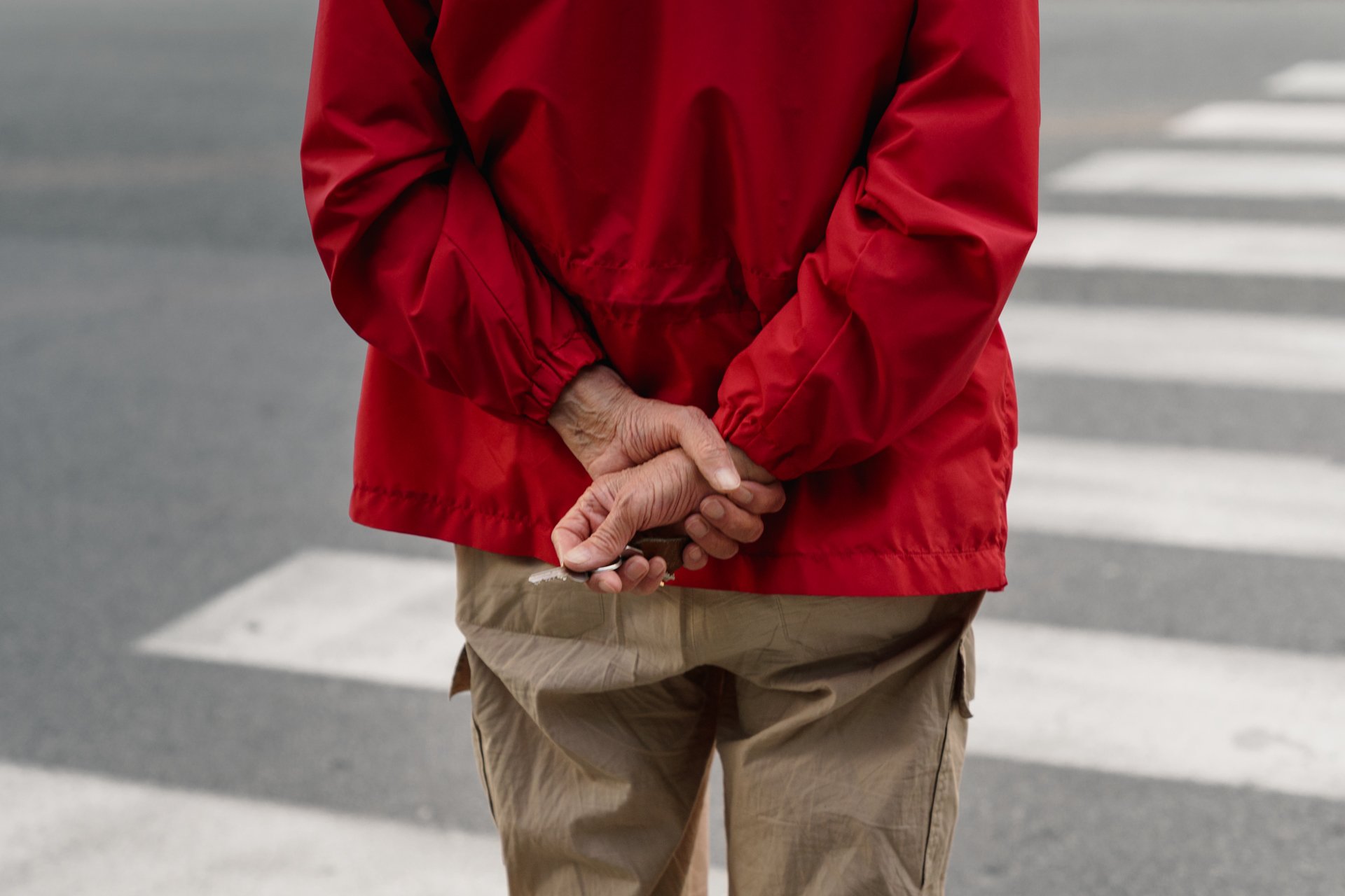 Una persona anziana con una giacca rossa e pantaloni beige si trova davanti a un attraversamento pedonale e tiene in mano un oggetto.