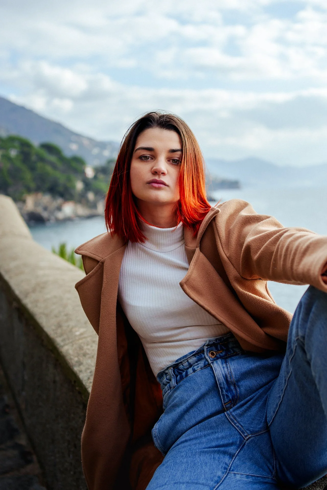 Una giovane donna con capelli rossi e vestiti casual si prende un selfie davanti a un paesaggio costiero con colline e mare in background.