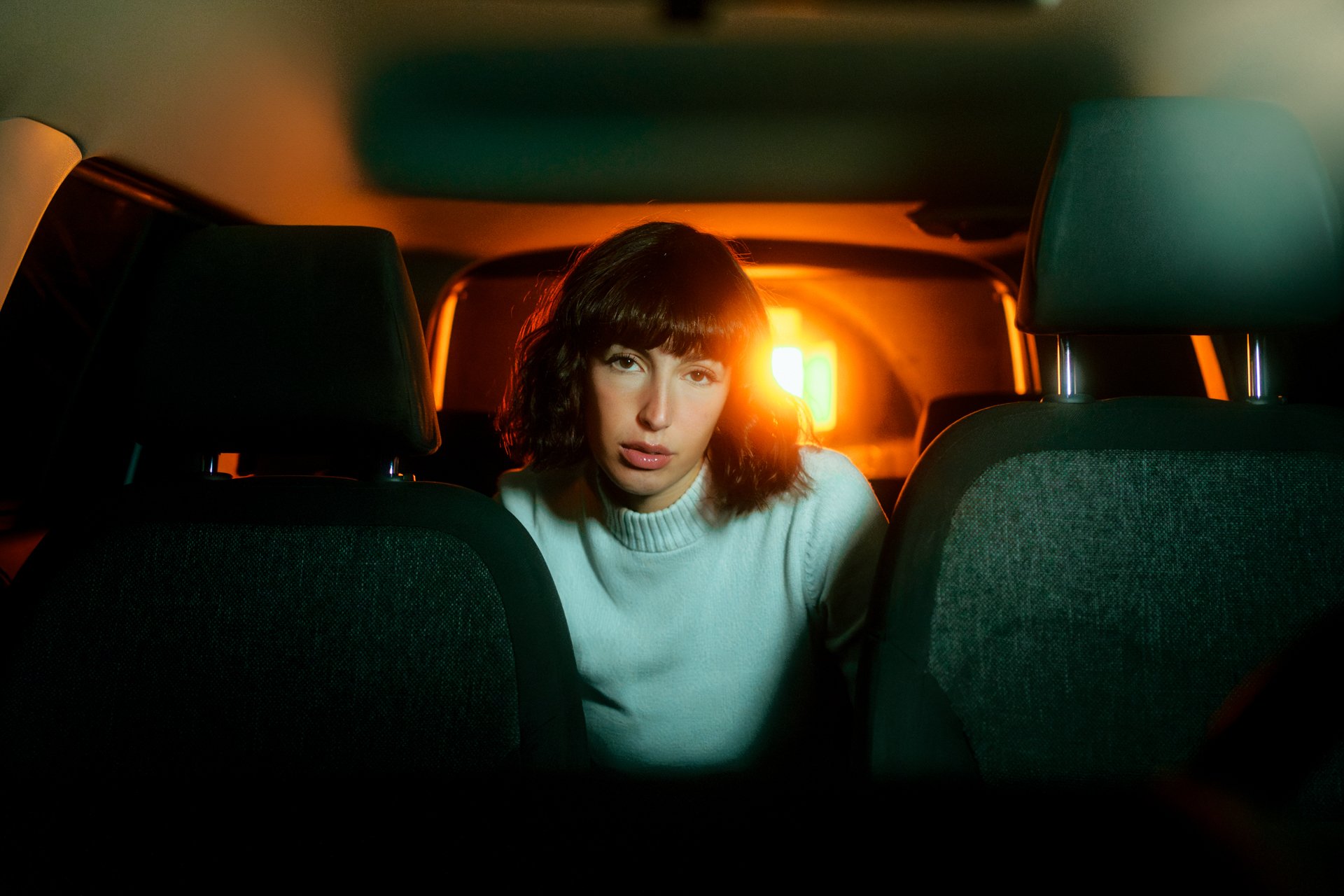 Ragazza seduta sul sedile di un'auto al crepuscolo, con luce arancione alle spalle, look casual a maglione bianco, espressione seria.
