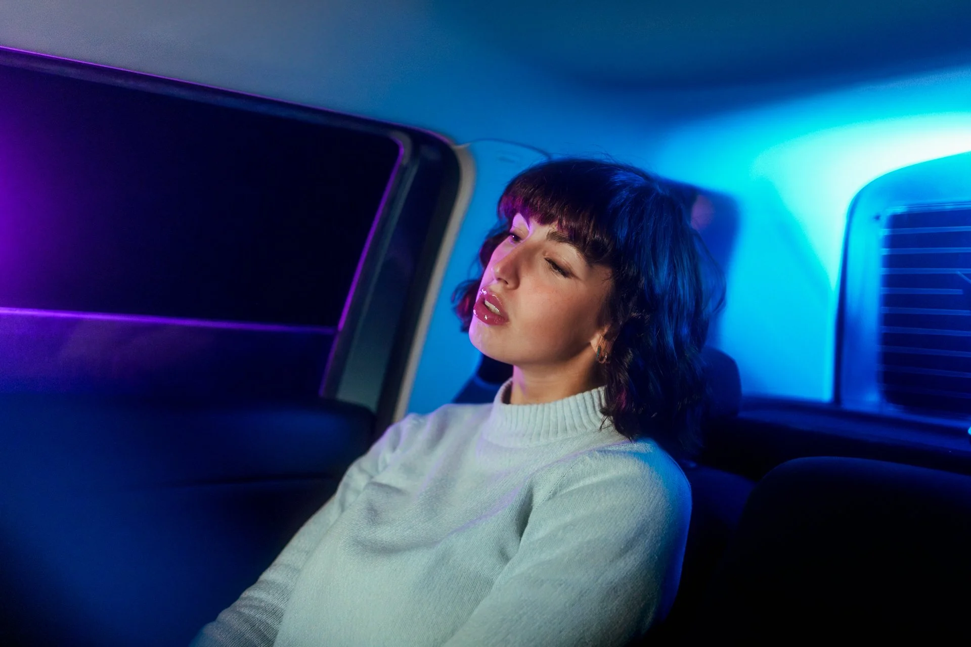 Una donna con capelli ricci scuri e cappelli, seduta in un'auto al buio con illuminazione colorata blu e viola.