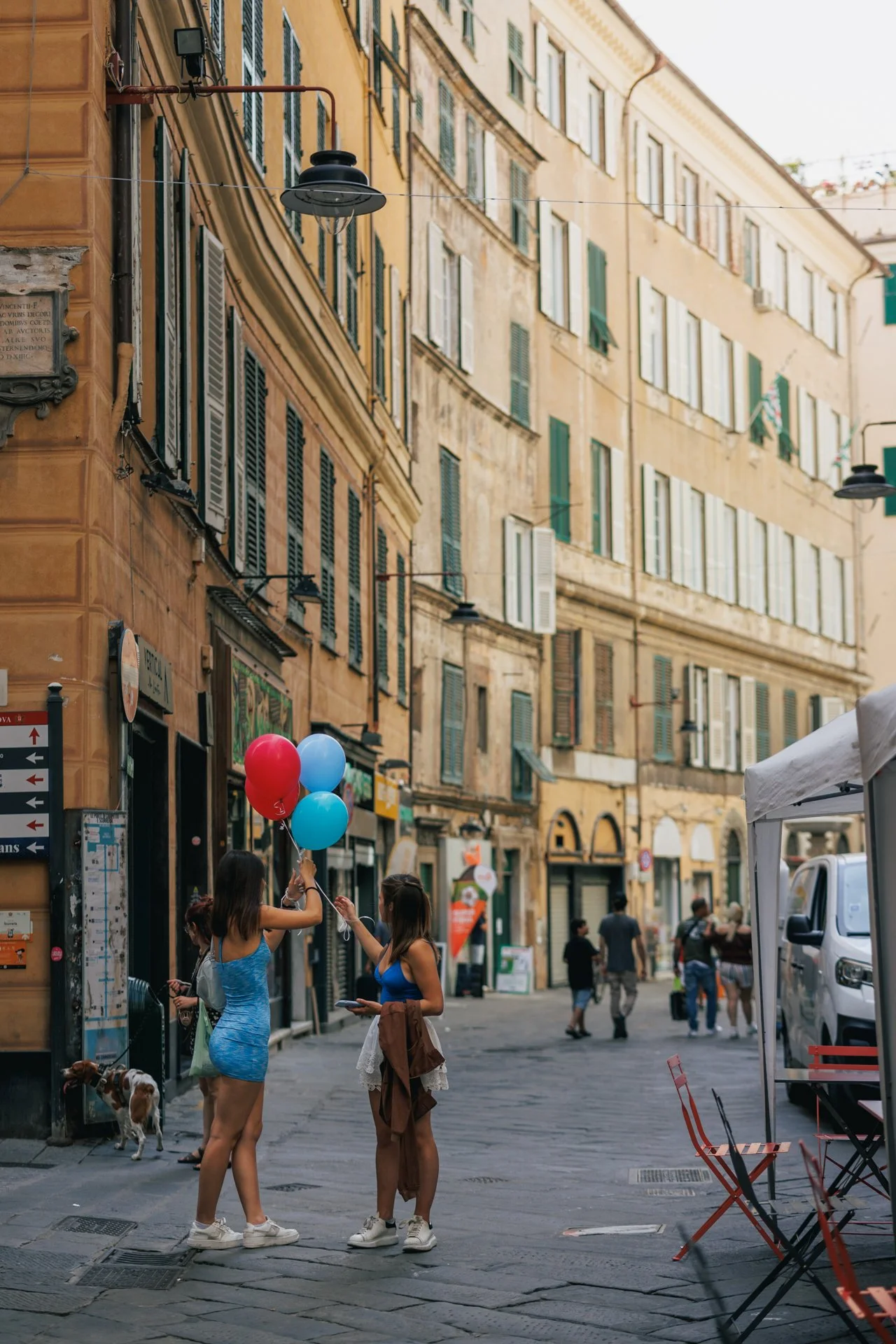 Due ragazze in abiti estivi con palloncini in una strada di città con edifici storici, alcuni cittadini camminano sullo sfondo, una donna con un cane al guinzaglio e sedie rosse di fronte a un'area all'aperto