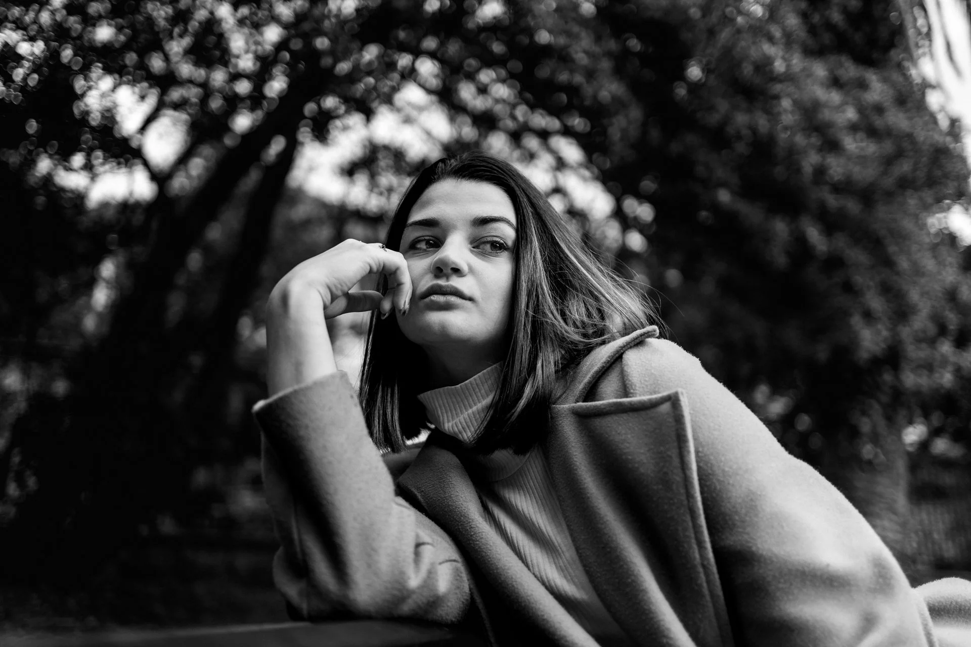 Ragazza con capelli lunghi e scuri, indossa una giacca, siede su una panchina in un parco, sfondo di alberi e natura, in bianco e nero.