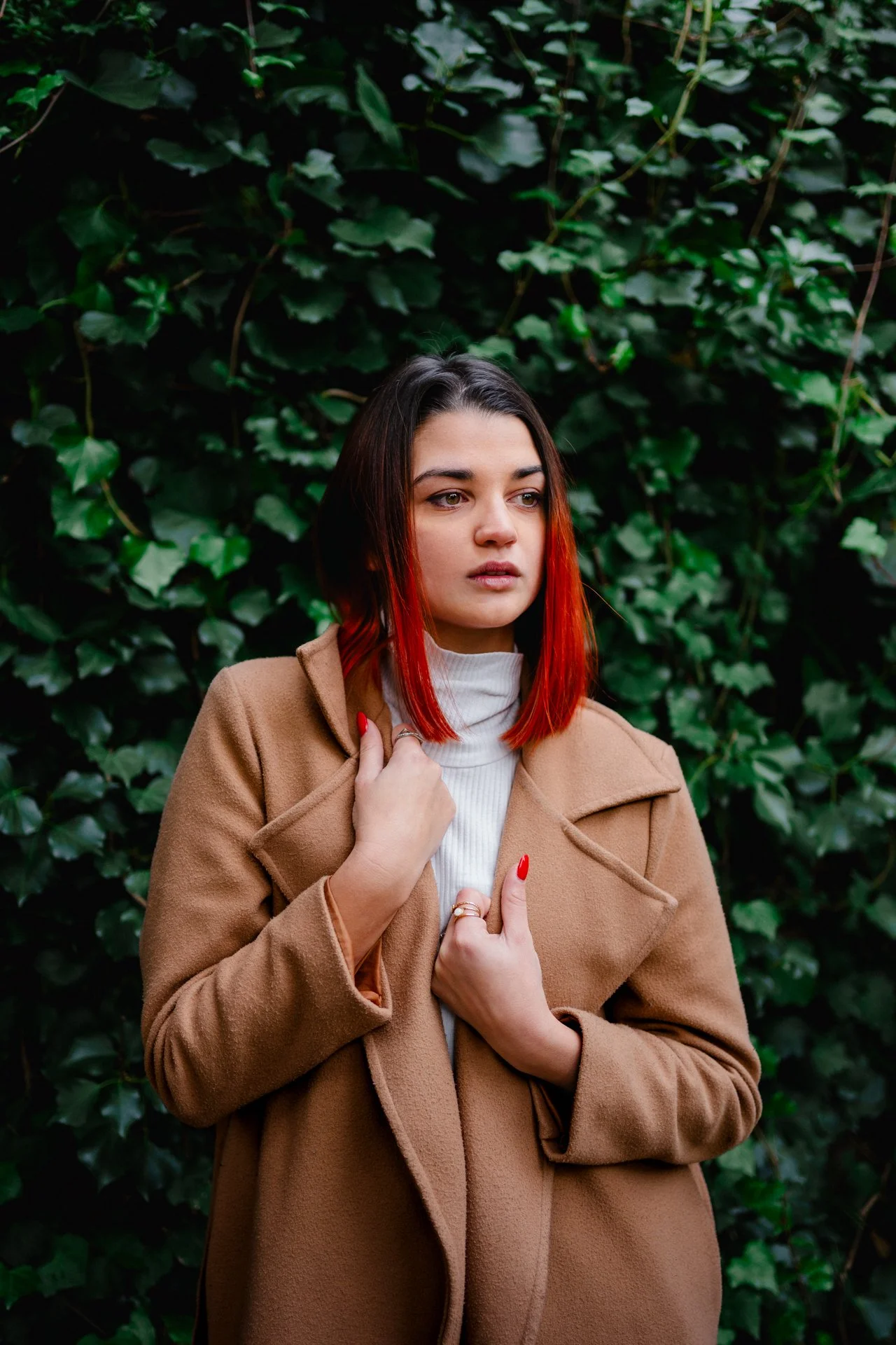 Ragazza con capelli castani e rossi, vestita con cappotto marrone e maglia bianca, posa davanti a una parete di foglie verdi