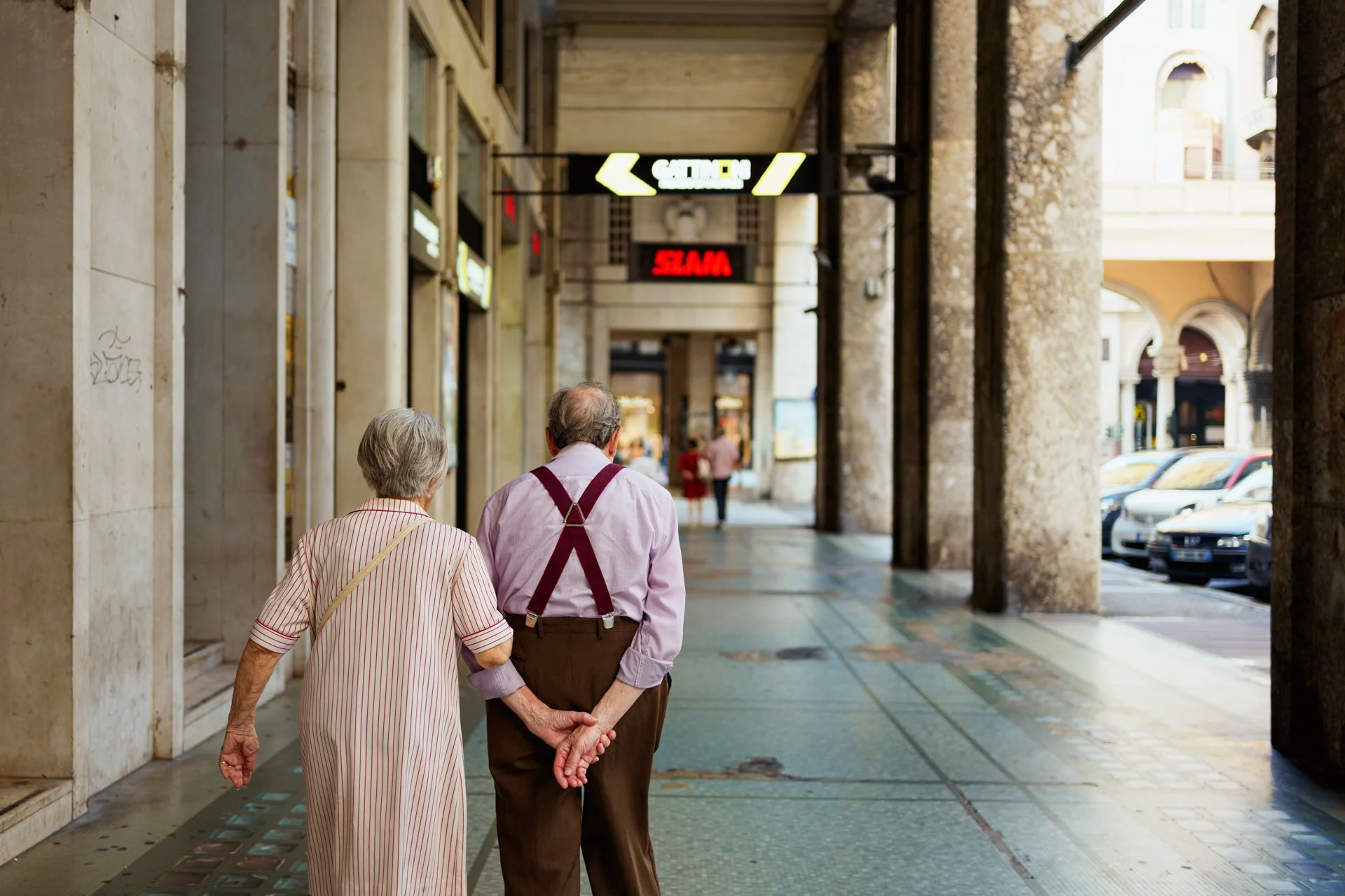 Due anziani passeggiano mano nella mano lungo un portico in un'area urbana, con negozi e auto parcheggiate visibili in fondo.