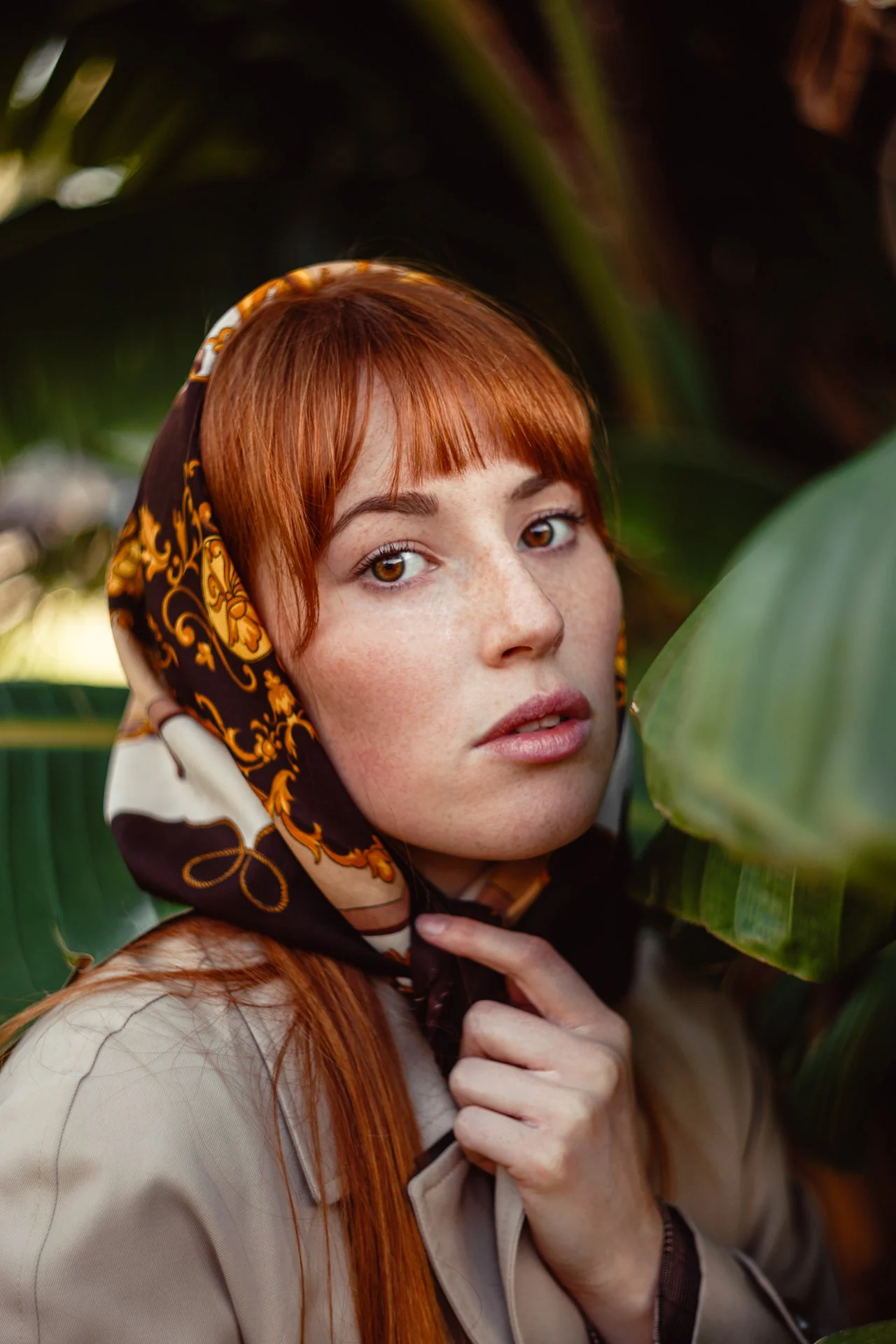 Ragazza con capelli rossi e lentiggini, indossa un foulard con disegni dorati e neri, si trova tra piante verdi.