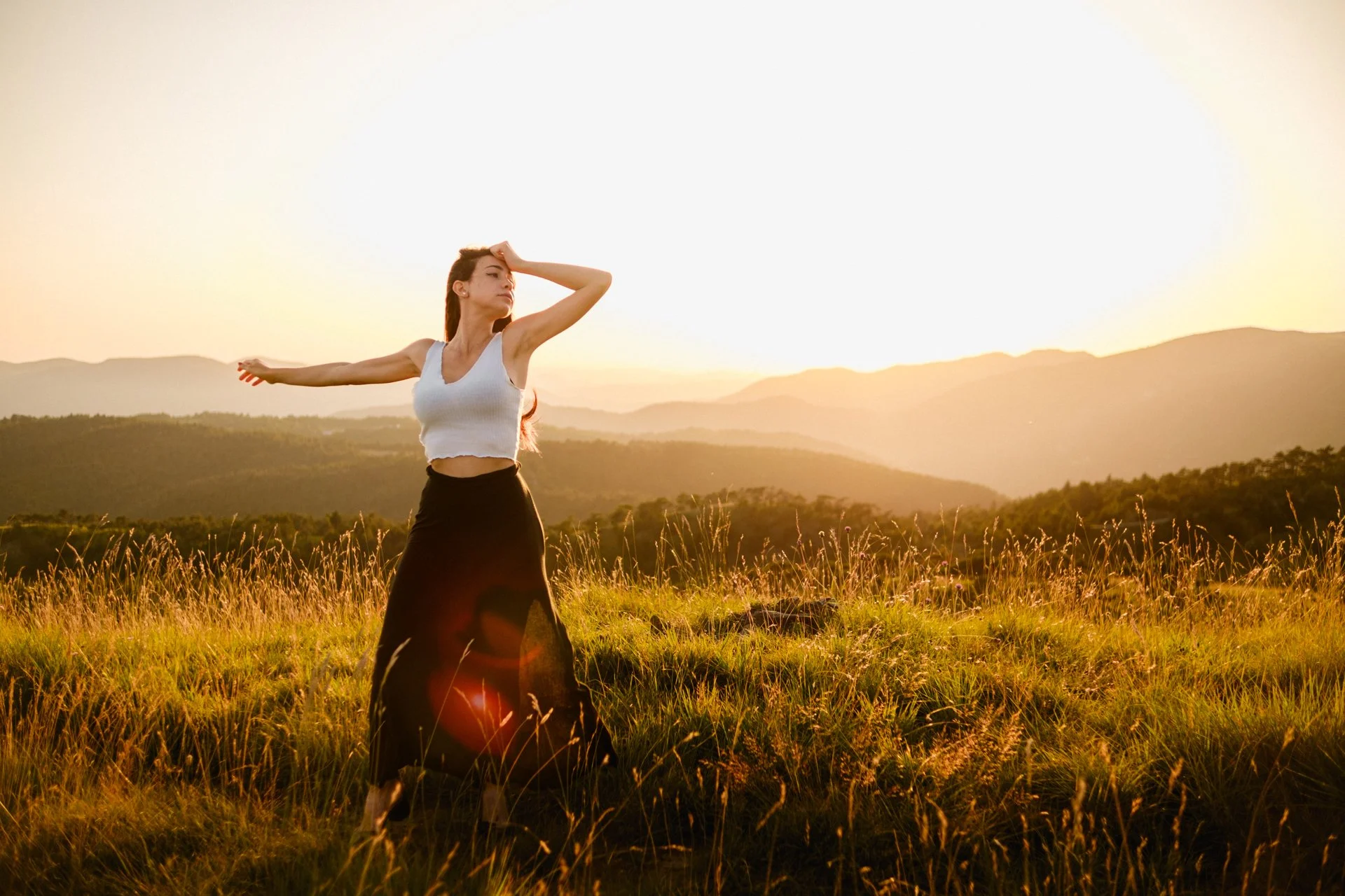 Una donna che si trova in un campo verde al tramonto, con le montagne sullo sfondo, indossa una maglietta bianca senza maniche e una gonna lunga nera.