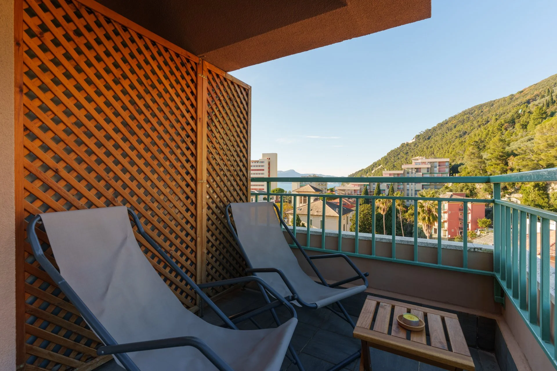 Balcone con due sedie a sdraio sullo sfondo di un paesaggio collinare con case e alberi, con pannelli in legno e ringhiera verde.