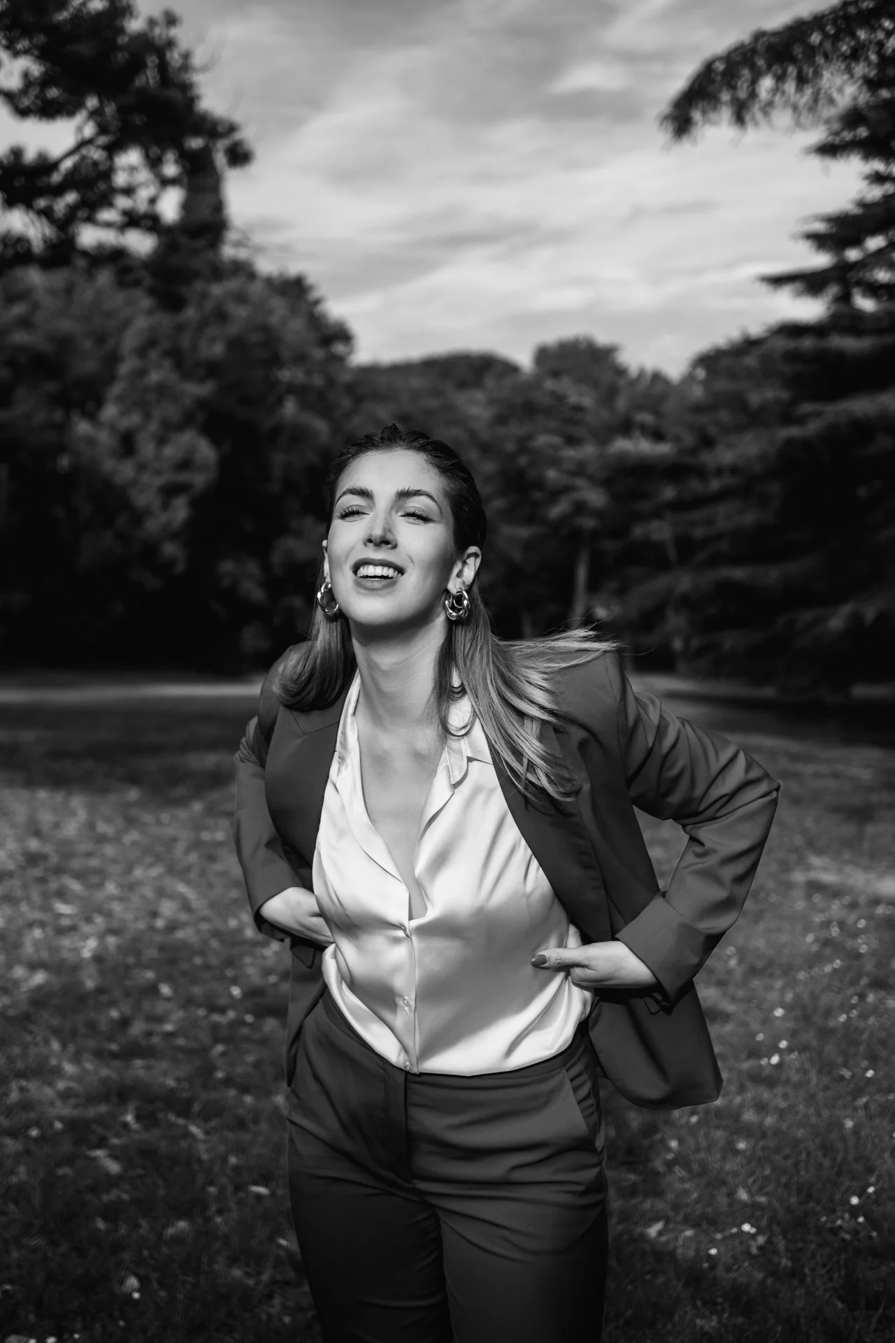 Una donna sorridente con i capelli sciolti indossa un abito elegante e una giacca casual in un parco, con alberi e cielo sullo sfondo in bianco e nero.