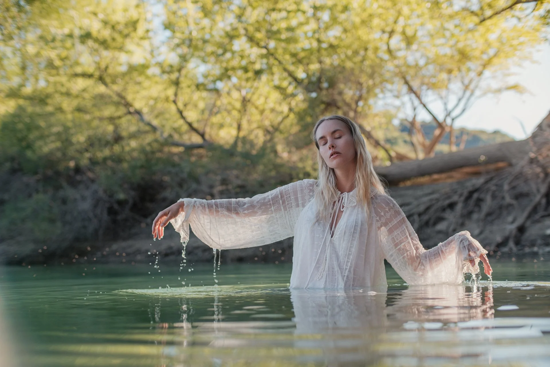 Una donna con capelli biondi e vestito bianco fa il bagno in un lago circondato da alberi verdi, con luce naturale e atmosfera tranquilla.