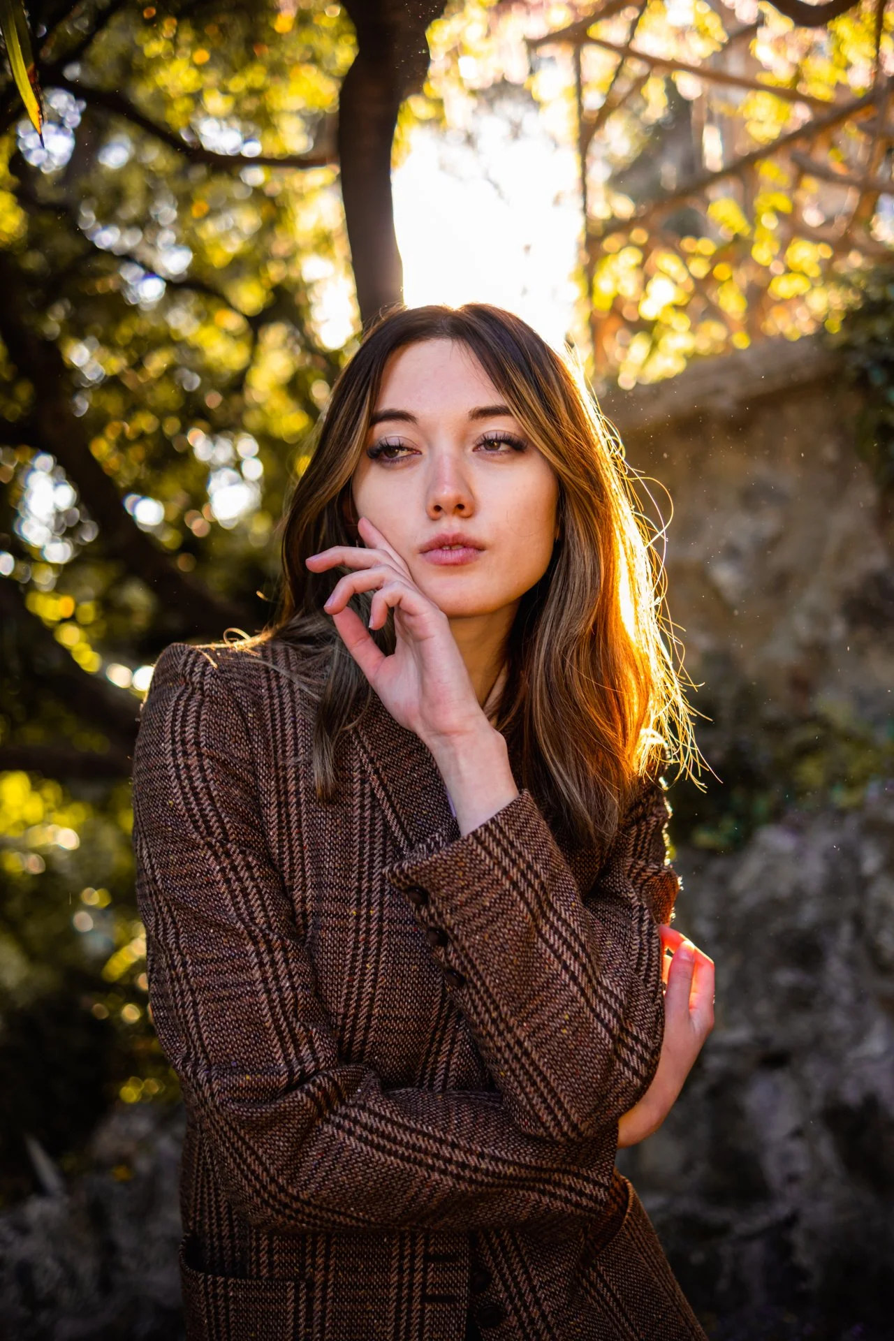 Ragazza con capelli castani e abito a quadri, posa con la mano vicino al viso in un contesto naturale con luce solare calda che filtra tra gli alberi.